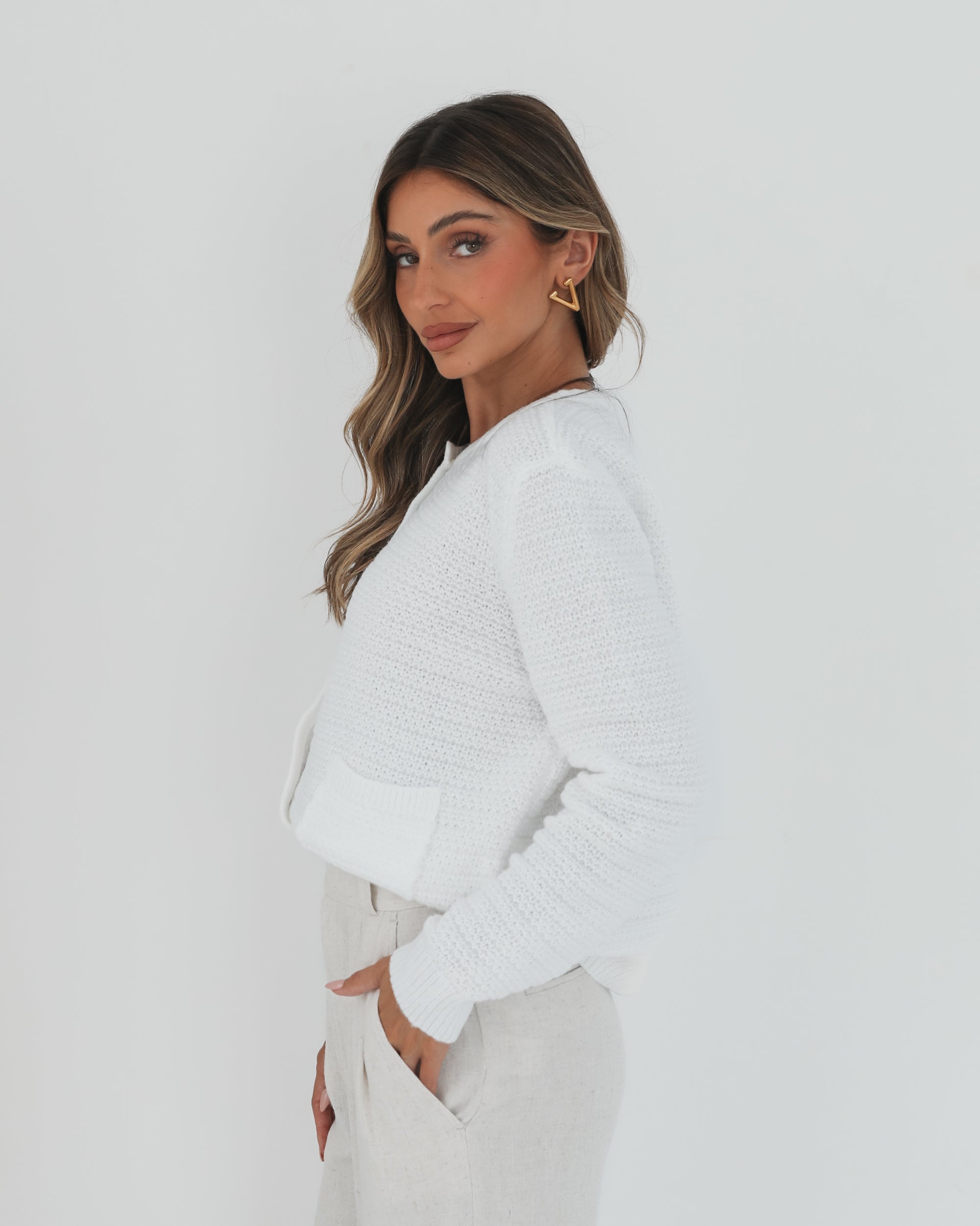 A woman wearing a white textured knit cardigan with front buttons and two small front pockets, paired with high-waisted light beige trousers. She stands against a plain white background, with one hand in her pocket and the other touching her hair.
