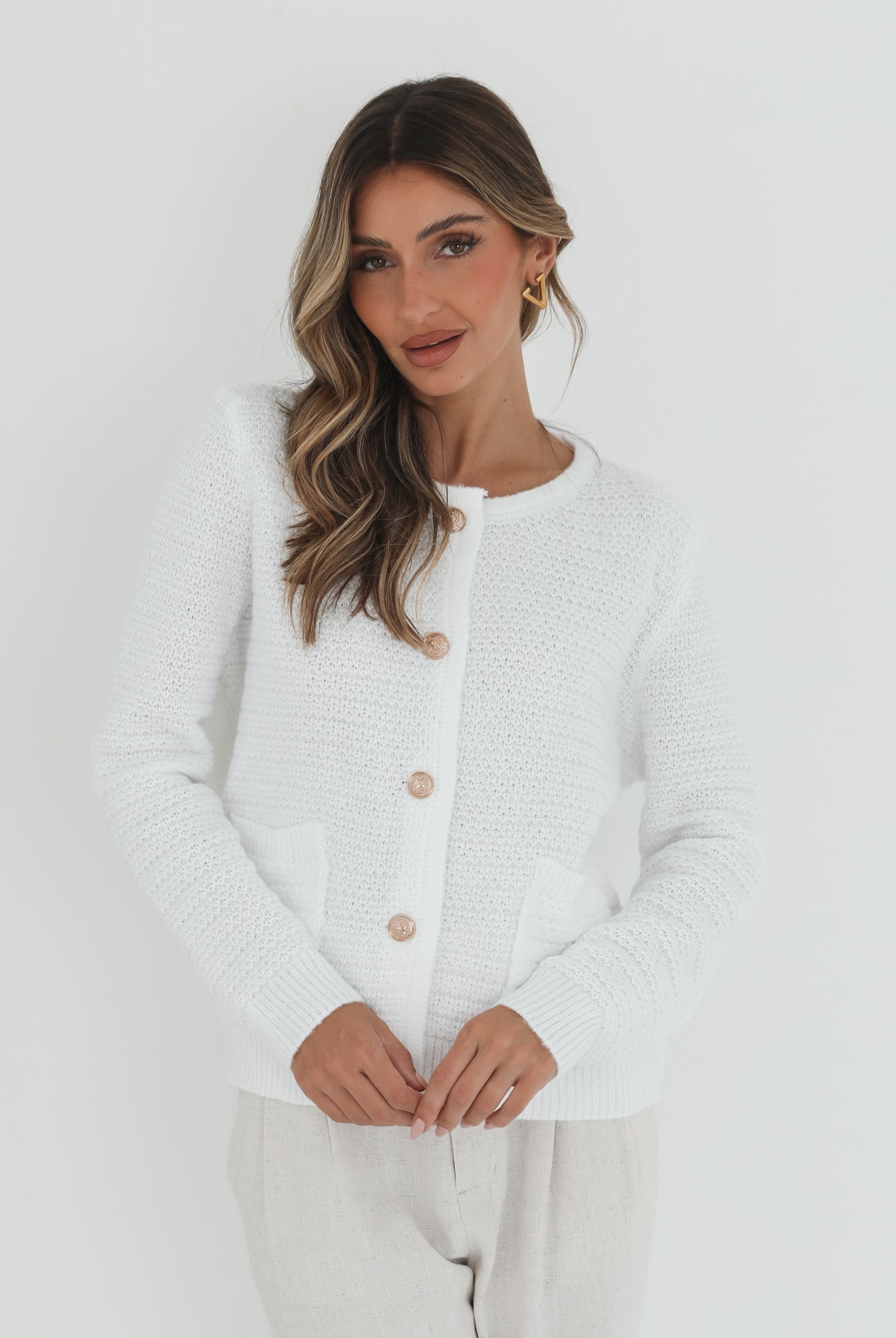 A woman wearing a white textured knit cardigan with front buttons and two small front pockets, paired with high-waisted light beige trousers. She stands against a plain white background, with one hand in her pocket and the other touching her hair.