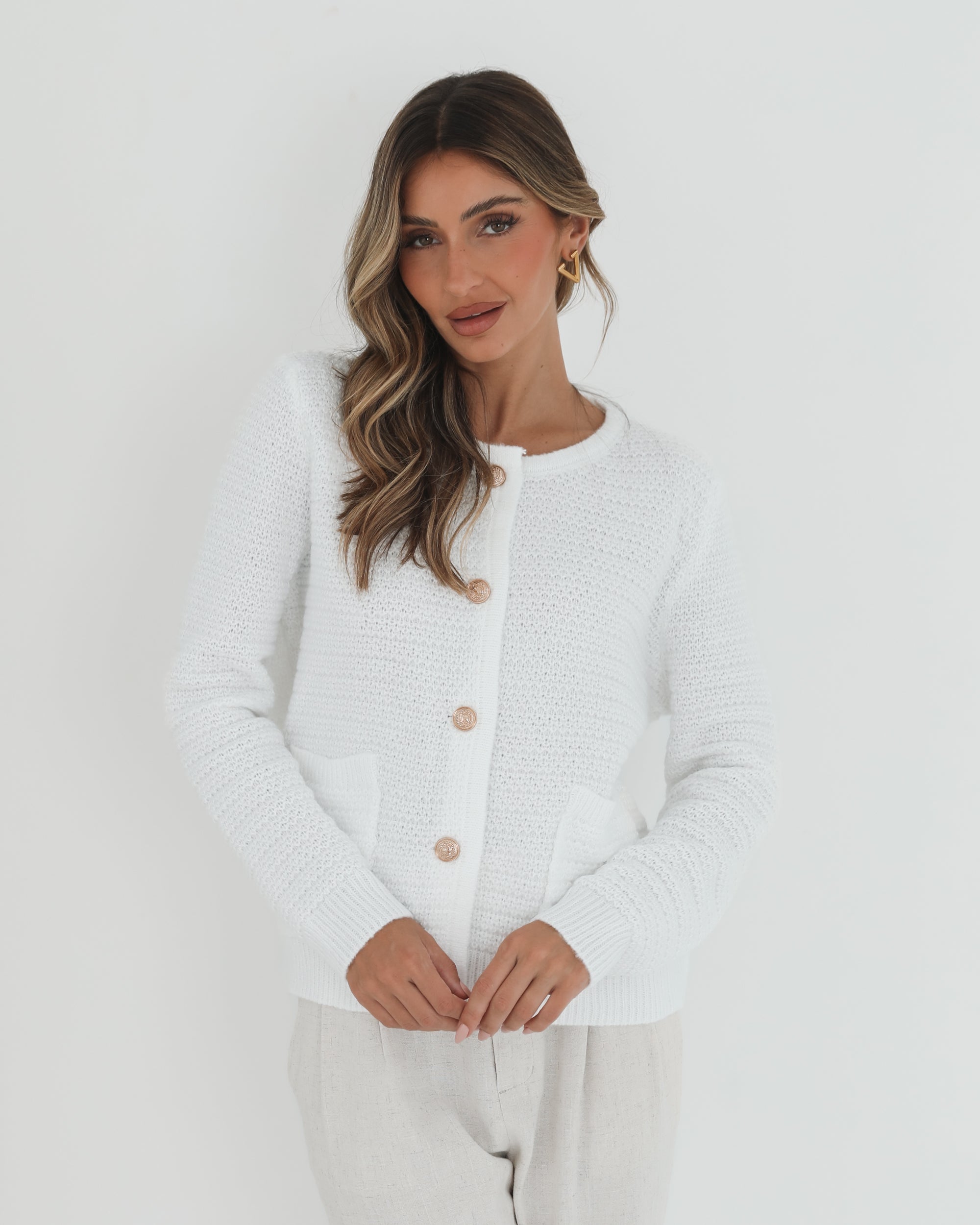 A woman wearing a white textured knit cardigan with front buttons and two small front pockets, paired with high-waisted light beige trousers. She stands against a plain white background, with one hand in her pocket and the other touching her hair.