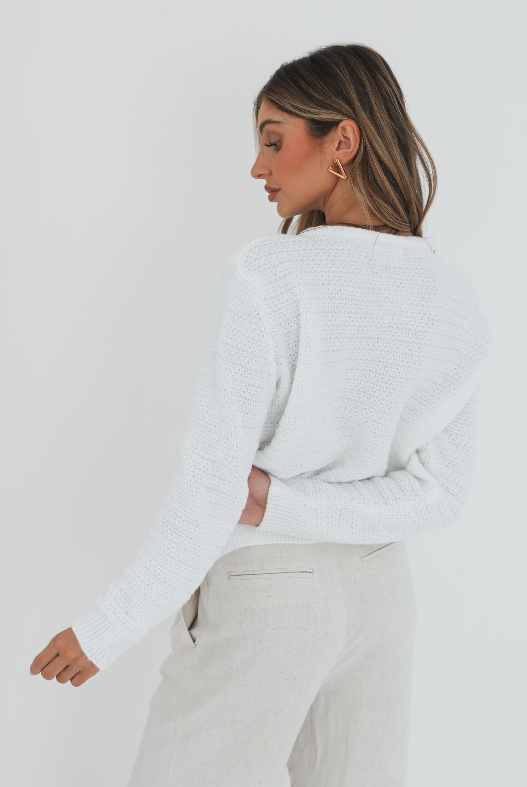 A woman wearing a white textured knit cardigan with front buttons and two small front pockets, paired with high-waisted light beige trousers. She stands against a plain white background, with one hand in her pocket and the other touching her hair.