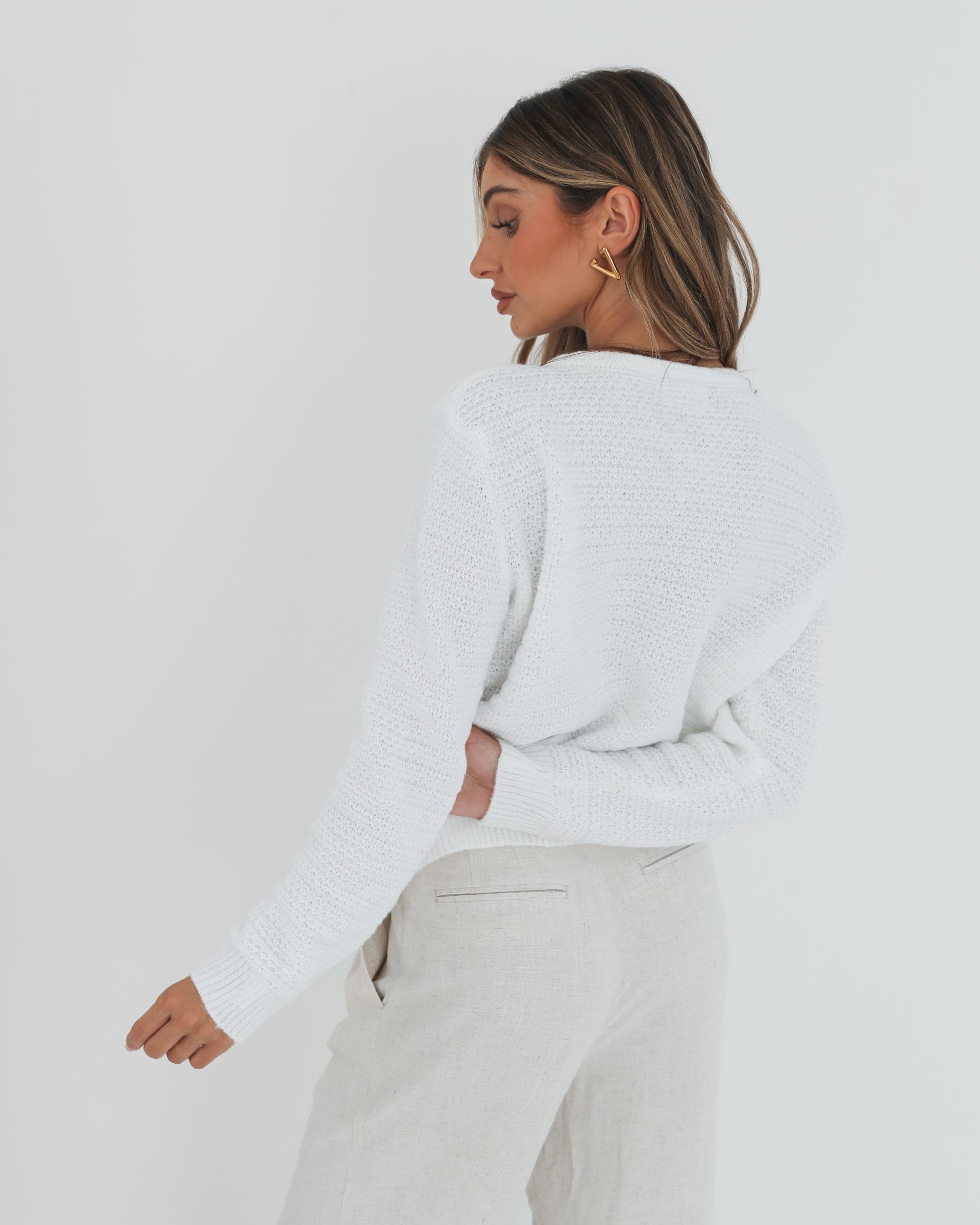 A woman wearing a white textured knit cardigan with front buttons and two small front pockets, paired with high-waisted light beige trousers. She stands against a plain white background, with one hand in her pocket and the other touching her hair.