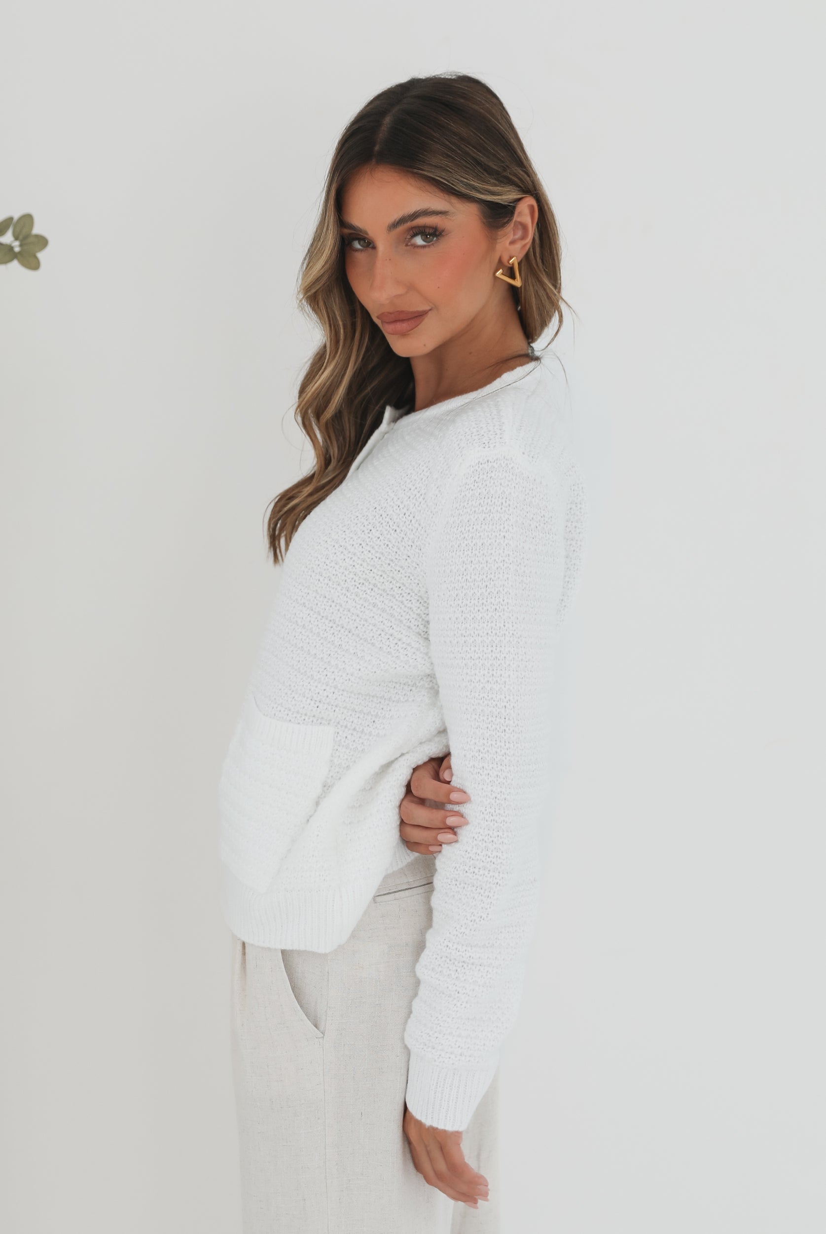 A woman wearing a white textured knit cardigan with front buttons and two small front pockets, paired with high-waisted light beige trousers. She stands against a plain white background, with one hand in her pocket and the other touching her hair.