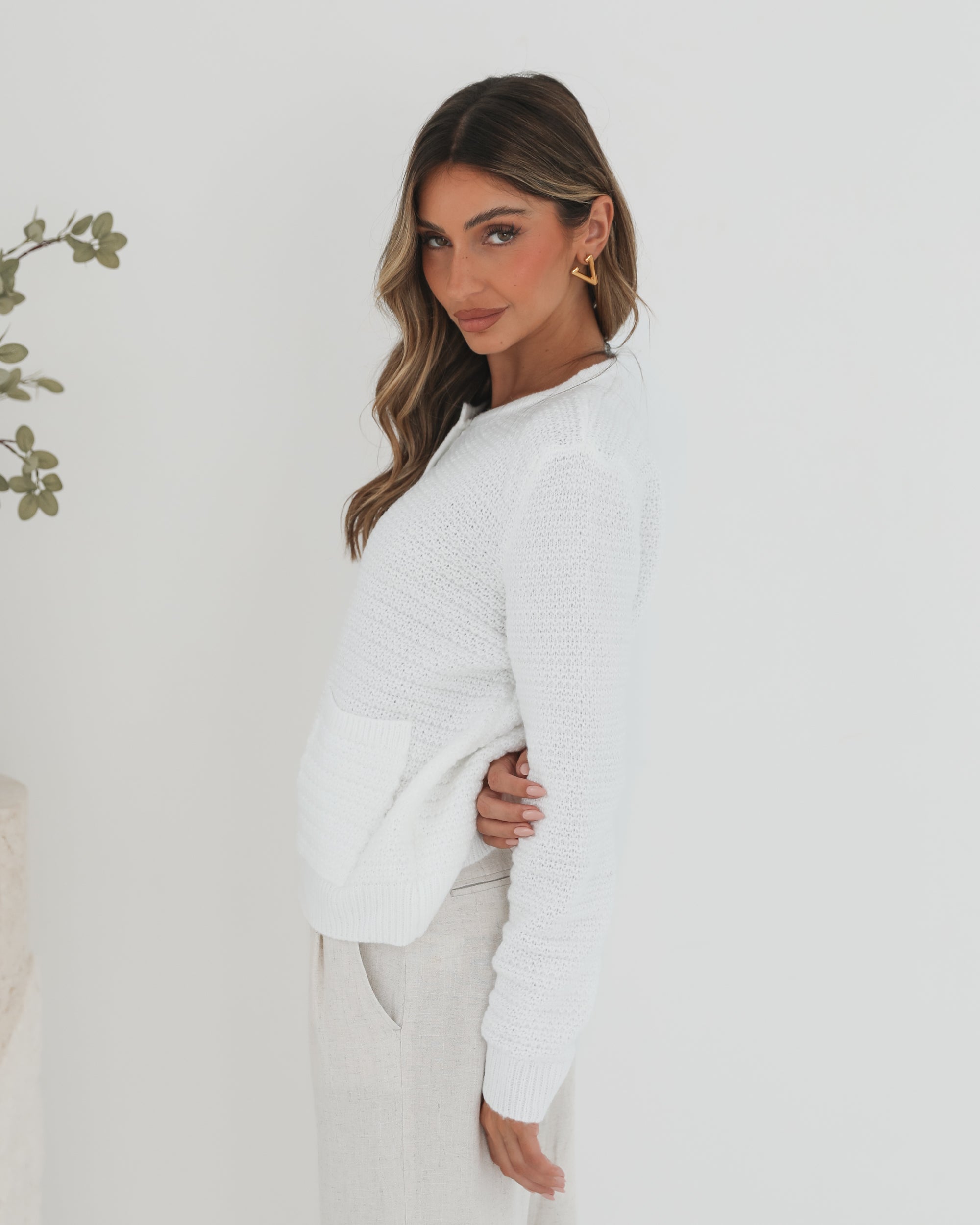 A woman wearing a white textured knit cardigan with front buttons and two small front pockets, paired with high-waisted light beige trousers. She stands against a plain white background, with one hand in her pocket and the other touching her hair.