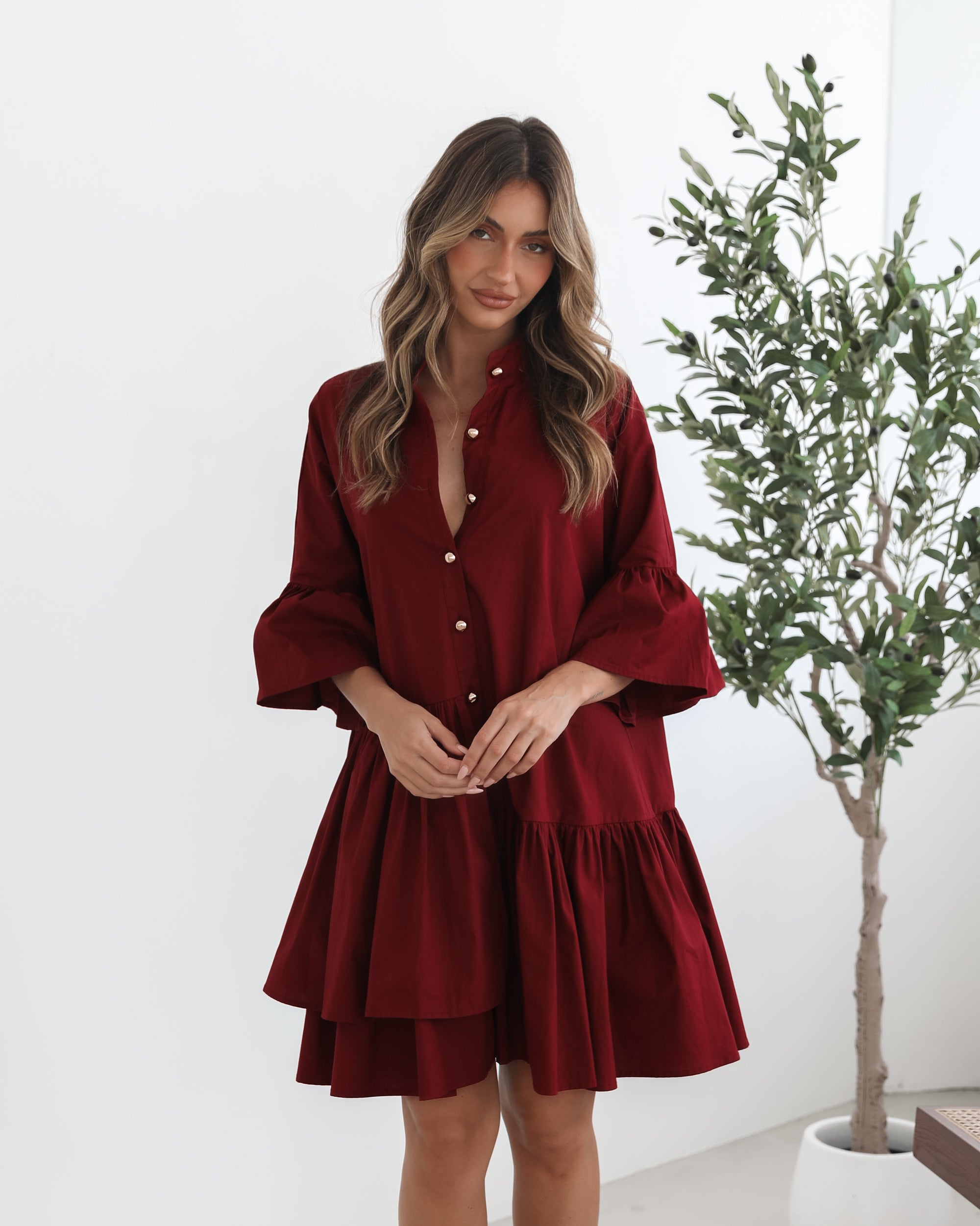 Smiling woman with long wavy hair wearing a loose-fitting, deep red button-up dress with ruffled tiers and three-quarter sleeves, paired with black ankle boots. She is standing indoors on a light-colored floor, holding the hem of her dress with one hand. A potted green plant and a wooden chair are visible in the background.