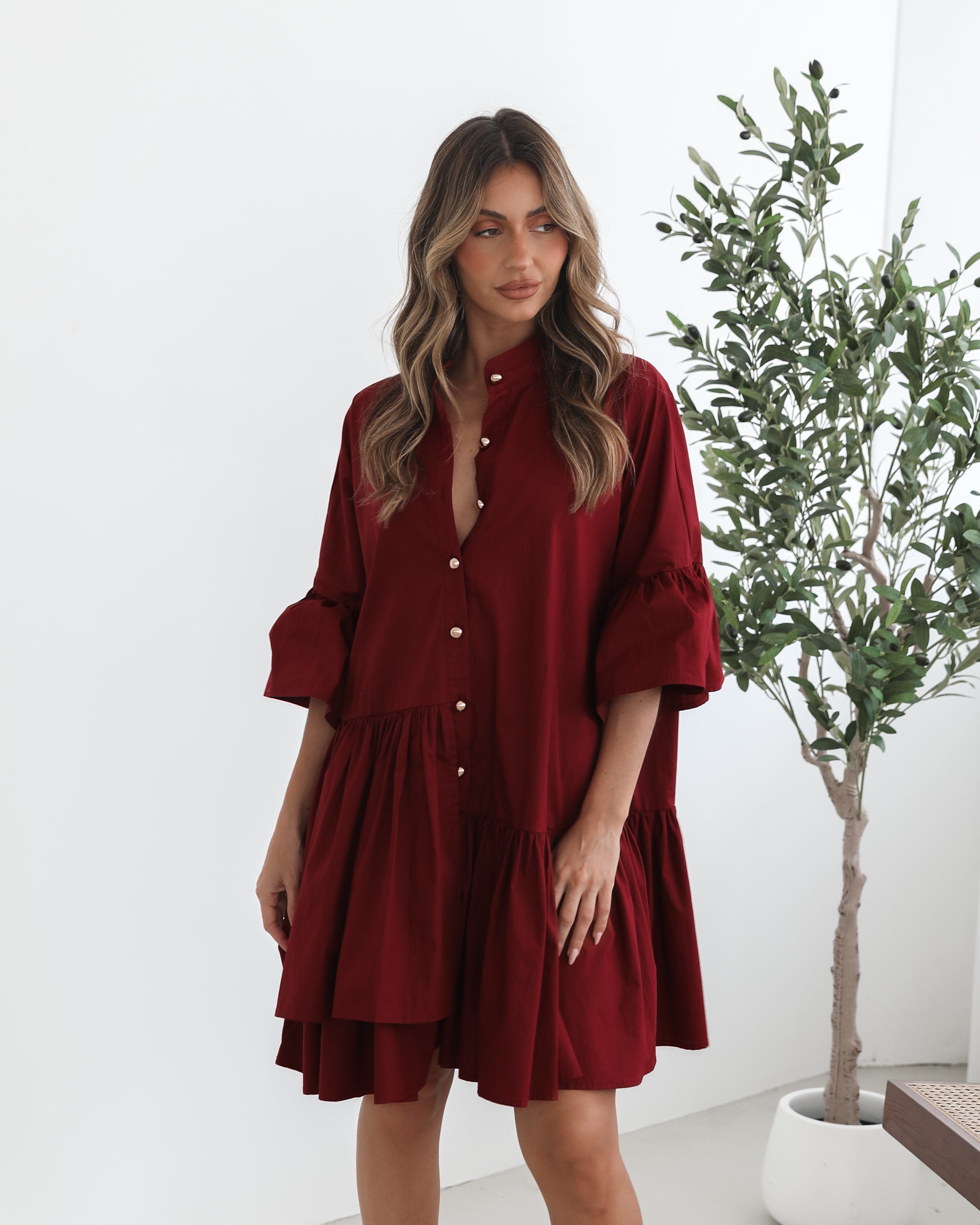 Smiling woman with long wavy hair wearing a loose-fitting, deep red button-up dress with ruffled tiers and three-quarter sleeves, paired with black ankle boots. She is standing indoors on a light-colored floor, holding the hem of her dress with one hand. A potted green plant and a wooden chair are visible in the background.