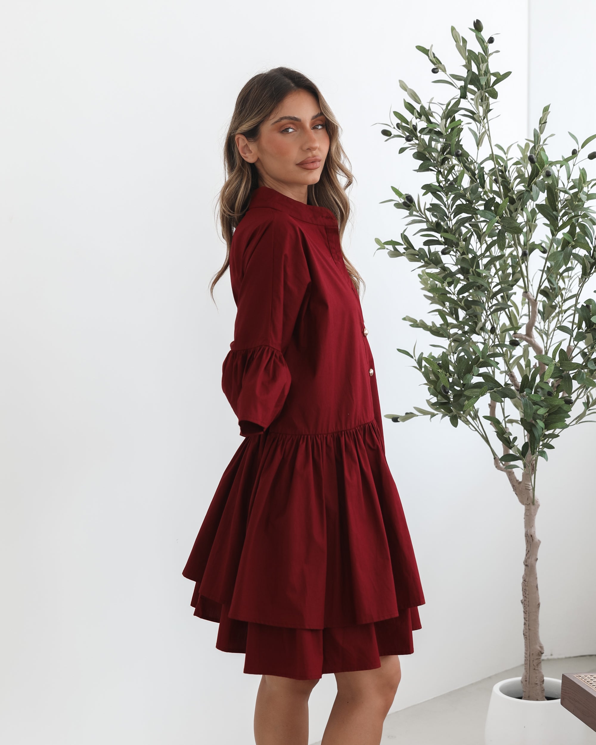 Smiling woman with long wavy hair wearing a loose-fitting, deep red button-up dress with ruffled tiers and three-quarter sleeves, paired with black ankle boots. She is standing indoors on a light-colored floor, holding the hem of her dress with one hand. A potted green plant and a wooden chair are visible in the background.