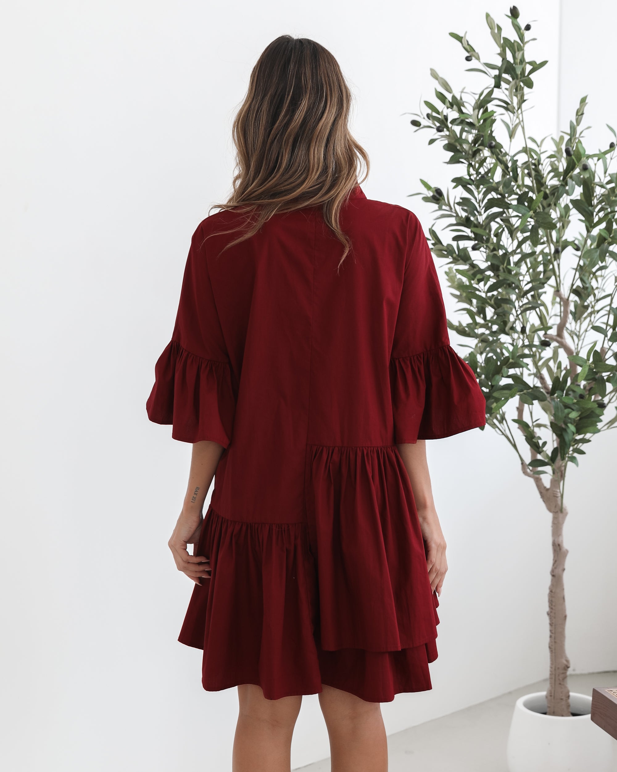 Smiling woman with long wavy hair wearing a loose-fitting, deep red button-up dress with ruffled tiers and three-quarter sleeves, paired with black ankle boots. She is standing indoors on a light-colored floor, holding the hem of her dress with one hand. A potted green plant and a wooden chair are visible in the background.