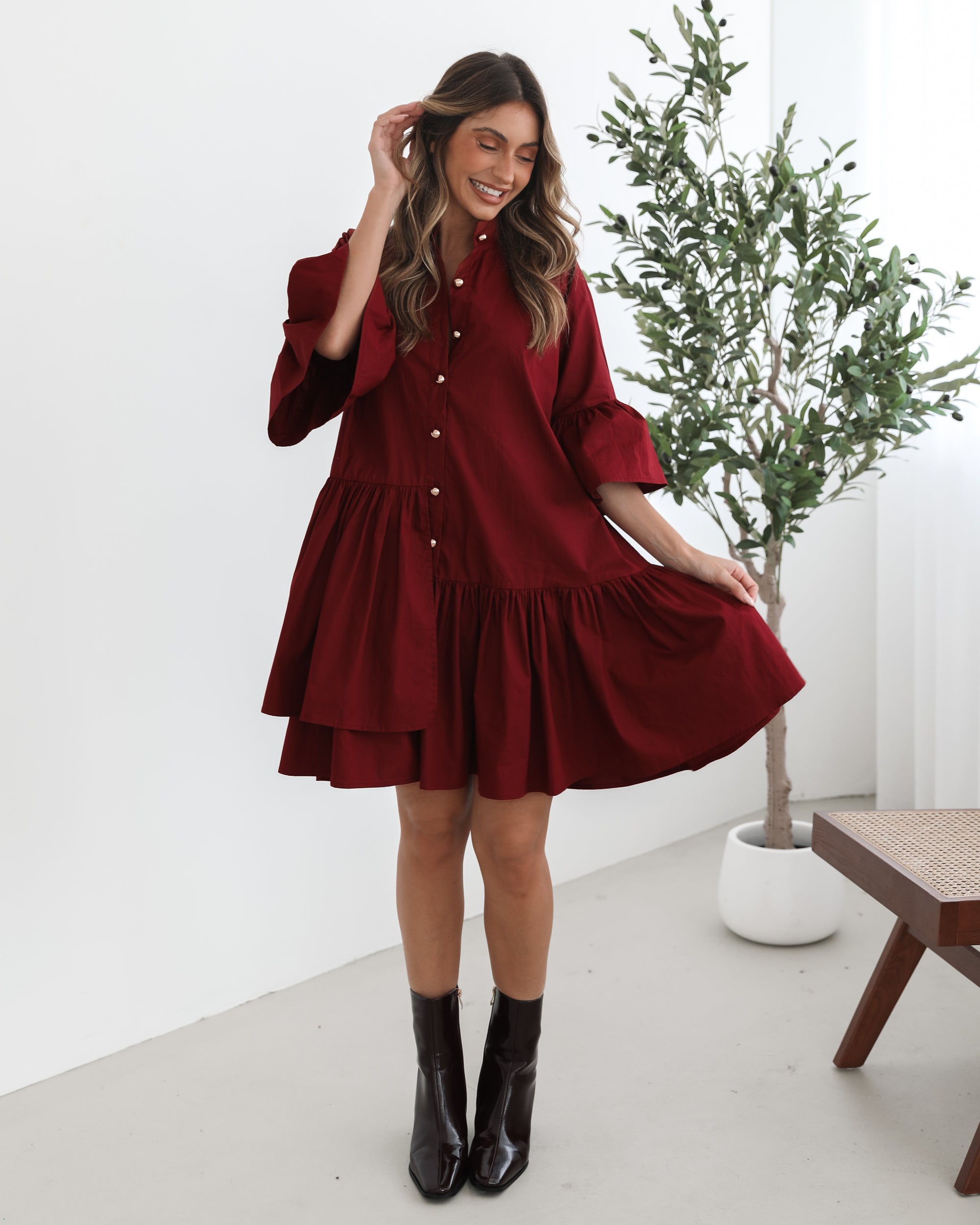 Smiling woman with long wavy hair wearing a loose-fitting, deep red button-up dress with ruffled tiers and three-quarter sleeves, paired with black ankle boots. She is standing indoors on a light-colored floor, holding the hem of her dress with one hand. A potted green plant and a wooden chair are visible in the background.