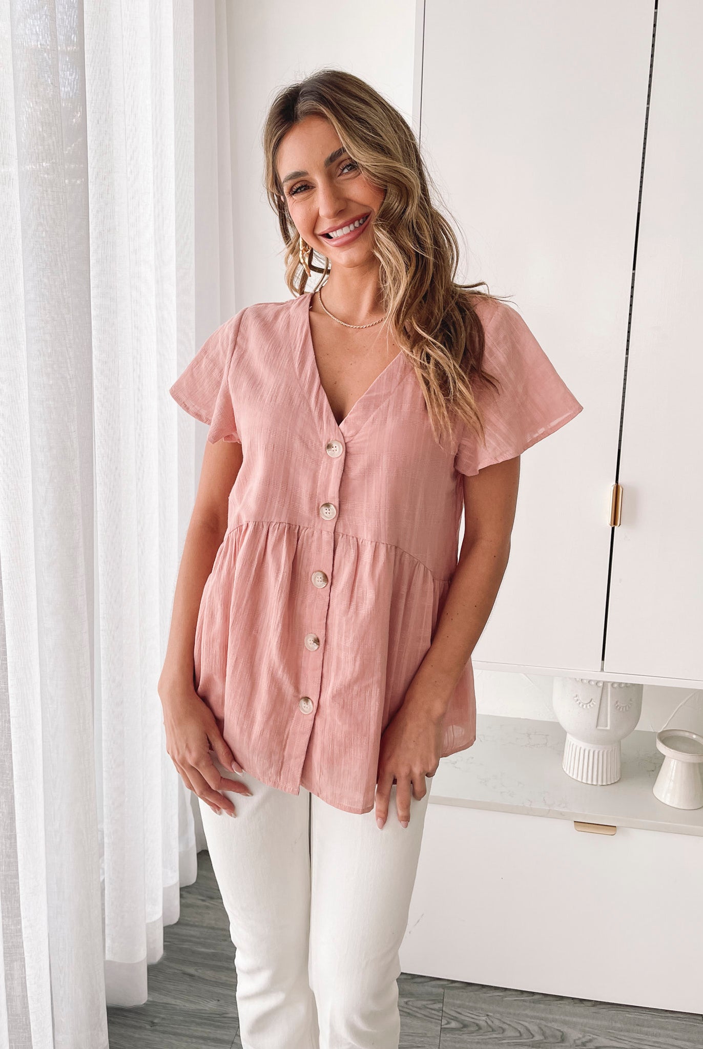 Smiling young woman with long blonde hair wearing a light pink button-up blouse and white shorts, standing indoors next to a white desk and wicker chair, with decorative art pieces in the background.