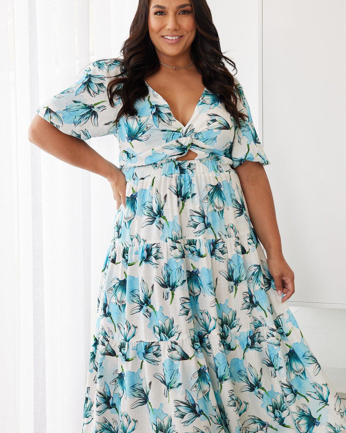 Woman wearing a white and blue floral midi dress with a front cut-out and ruched detailing, featuring elbow-length sleeves and a flowy skirt, standing indoors and smiling.