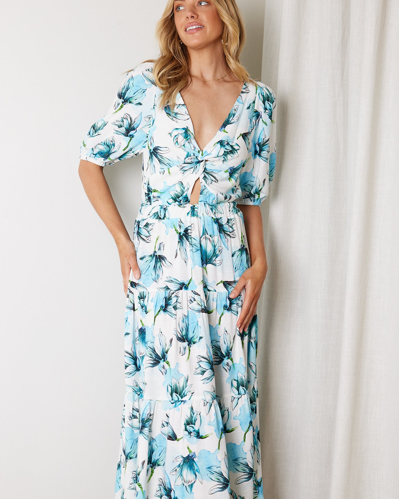 Woman wearing a white and blue floral midi dress with a front cut-out and ruched detailing, featuring elbow-length sleeves and a flowy skirt, standing indoors and smiling.
