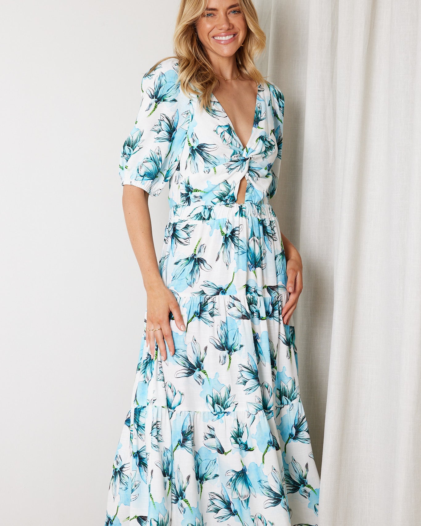 Woman wearing a white and blue floral midi dress with a front cut-out and ruched detailing, featuring elbow-length sleeves and a flowy skirt, standing indoors and smiling.