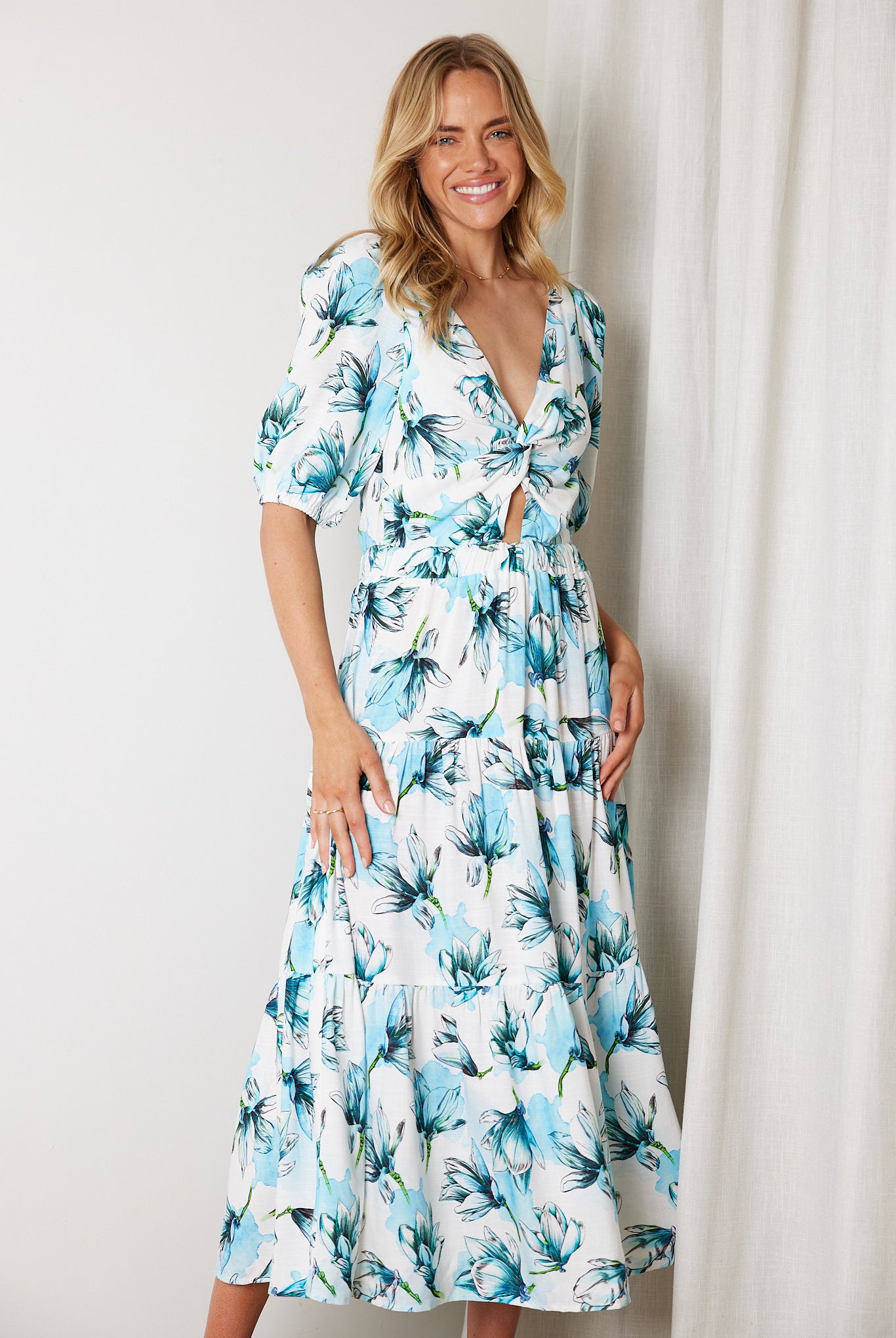 Woman wearing a white and blue floral midi dress with a front cut-out and ruched detailing, featuring elbow-length sleeves and a flowy skirt, standing indoors and smiling.