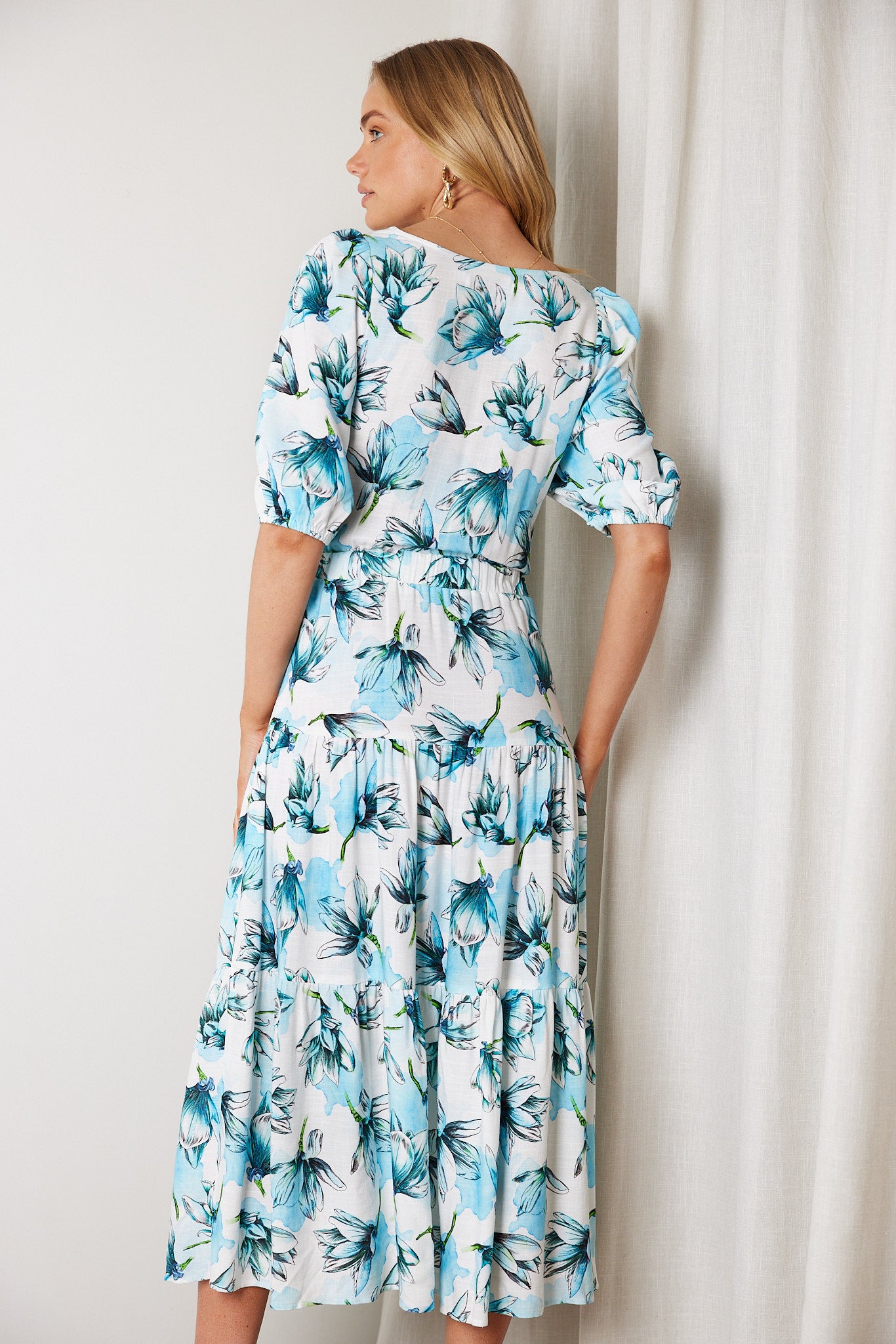 Woman wearing a white and blue floral midi dress with a front cut-out and ruched detailing, featuring elbow-length sleeves and a flowy skirt, standing indoors and smiling.
