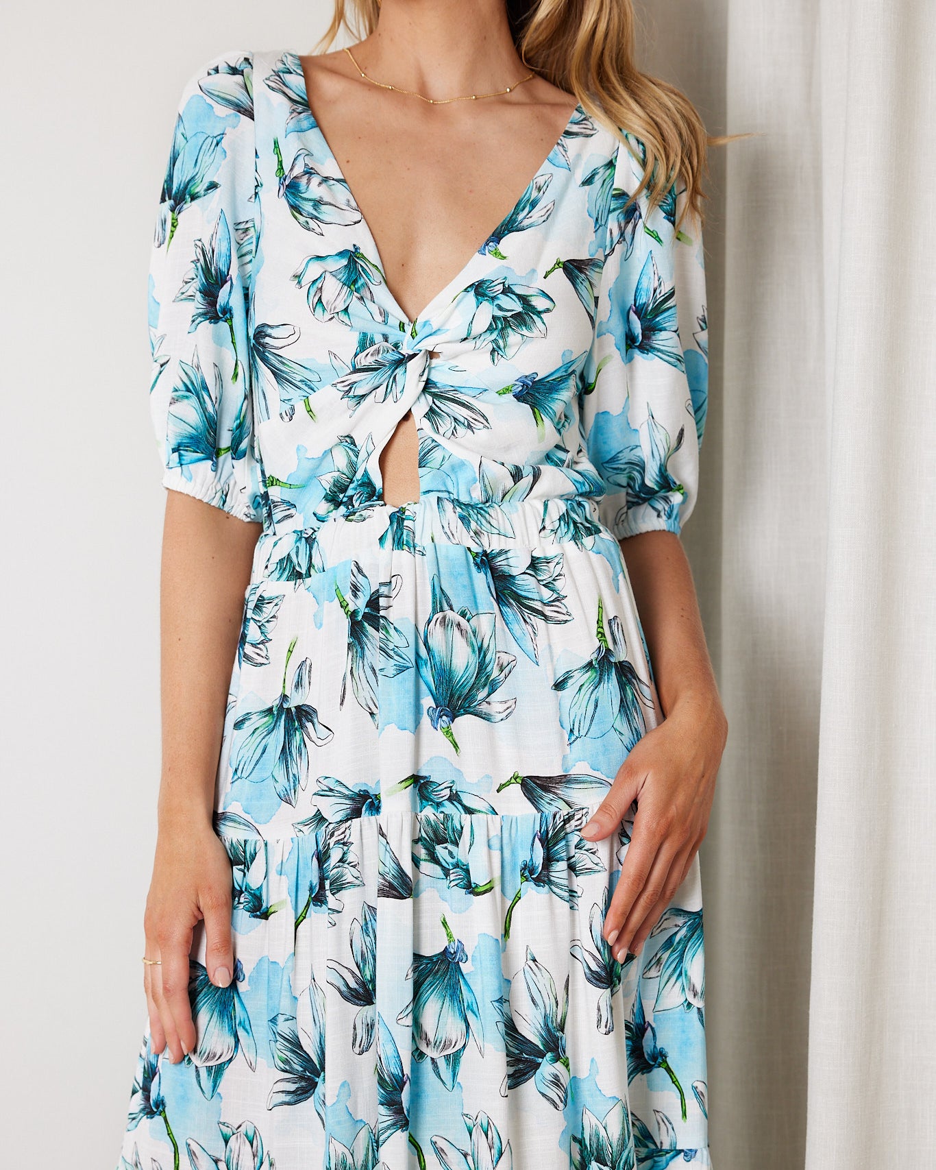 Woman wearing a white and blue floral midi dress with a front cut-out and ruched detailing, featuring elbow-length sleeves and a flowy skirt, standing indoors and smiling.