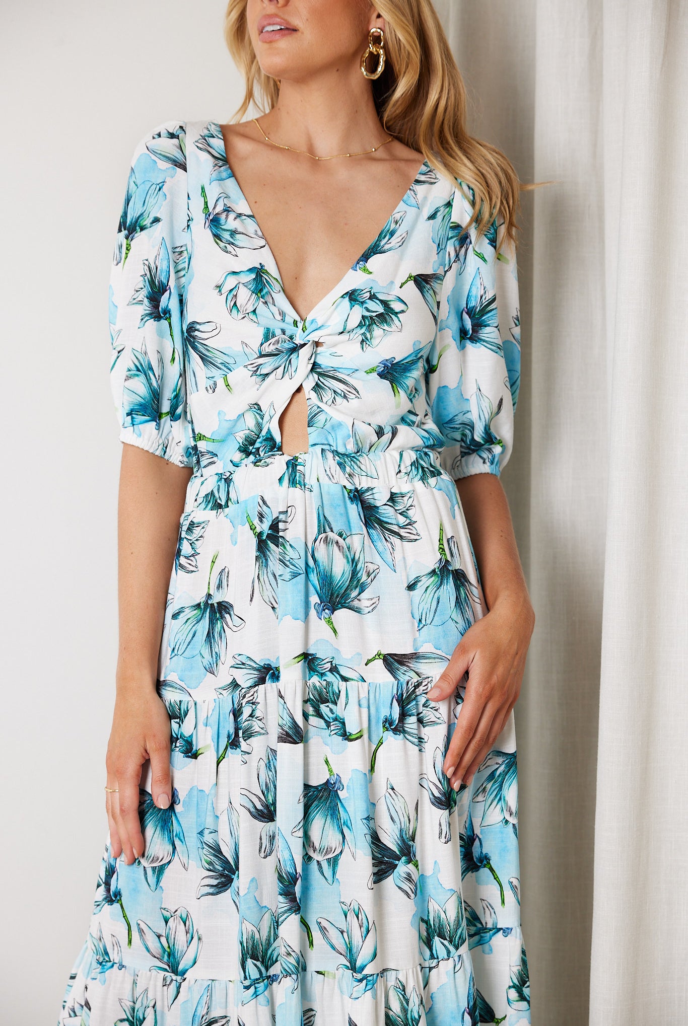 Woman wearing a white and blue floral midi dress with a front cut-out and ruched detailing, featuring elbow-length sleeves and a flowy skirt, standing indoors and smiling.
