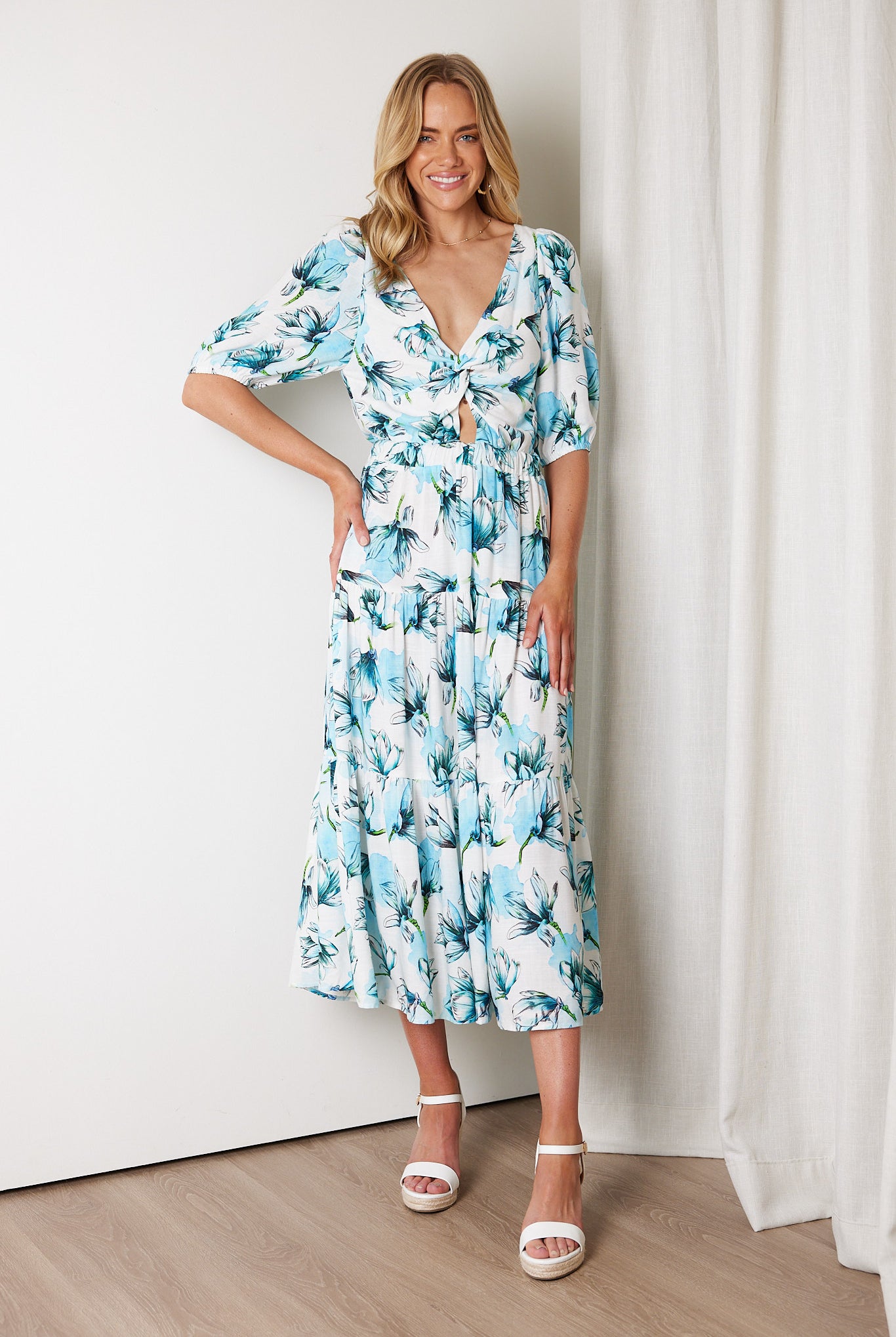 Woman wearing a white and blue floral midi dress with a front cut-out and ruched detailing, featuring elbow-length sleeves and a flowy skirt, standing indoors and smiling.