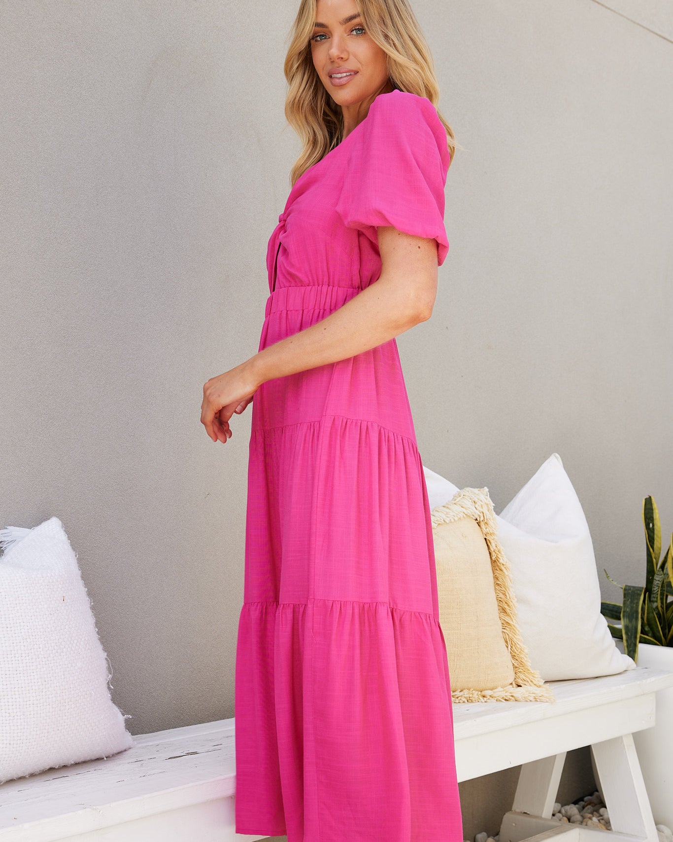 Woman wearing a bright pink short-sleeve midi dress with a front twist and cut-out detail, standing outdoors on white pebbles beside a white bench and potted plant.