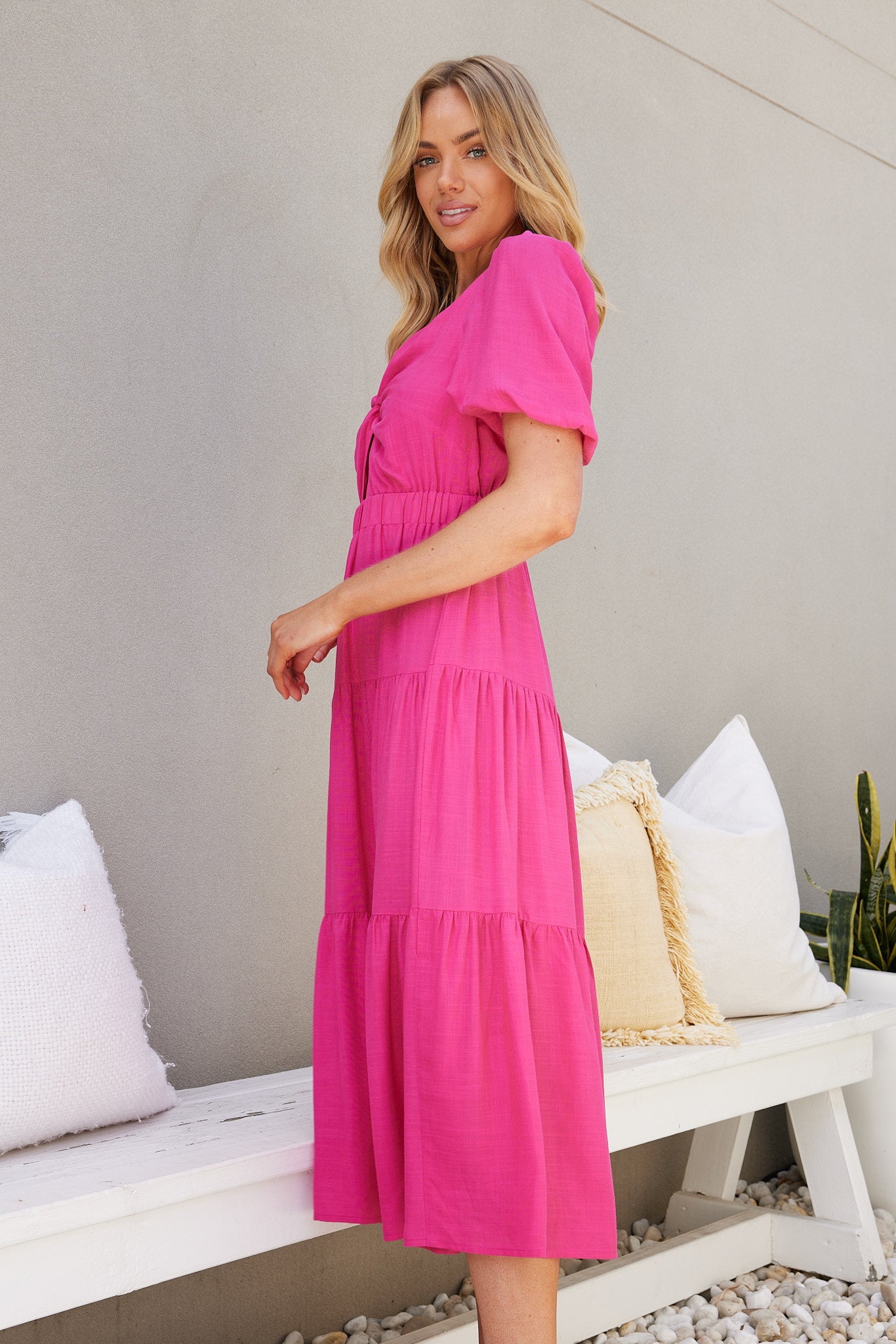 Woman wearing a bright pink short-sleeve midi dress with a front twist and cut-out detail, standing outdoors on white pebbles beside a white bench and potted plant.