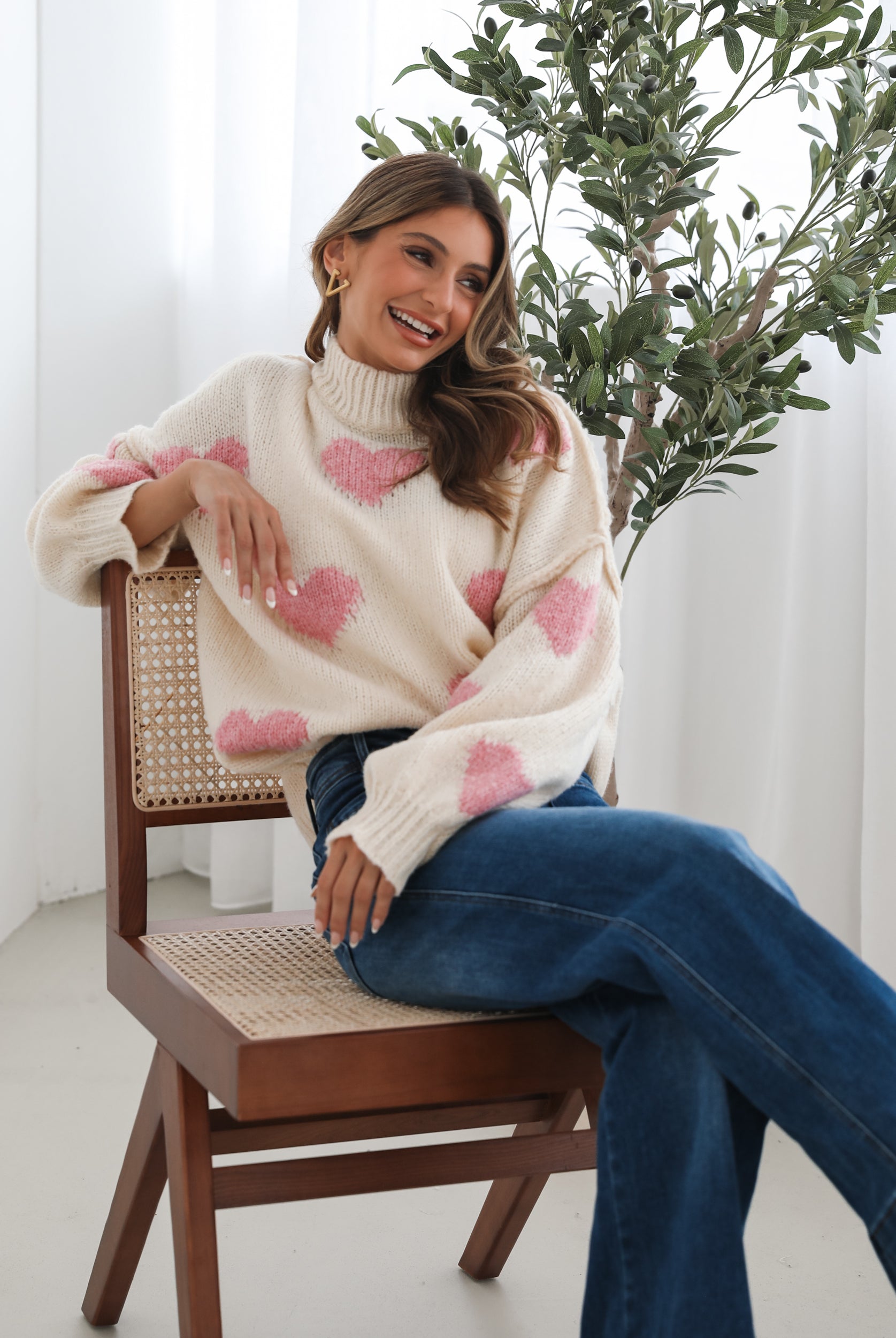 A woman sits smiling on a wooden chair with a woven back and seat. She is wearing a cozy cream-colored sweater with pink heart patterns and dark blue wide-leg jeans. Her long, wavy hair is styled to one side, and she accessorizes with gold triangle-shaped earrings. A tall green leafy plant stands in the background against white curtains, adding a fresh, modern touch to the bright, minimal setting