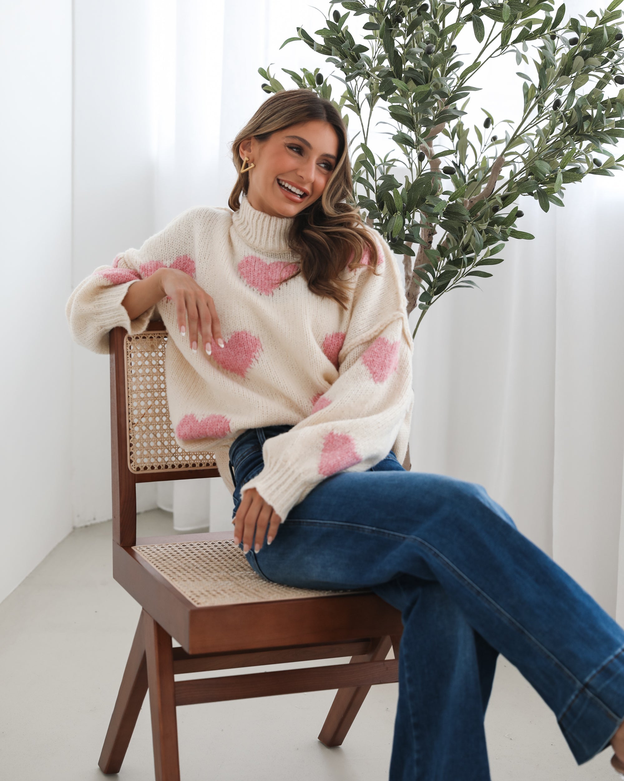 A woman sits smiling on a wooden chair with a woven back and seat. She is wearing a cozy cream-colored sweater with pink heart patterns and dark blue wide-leg jeans. Her long, wavy hair is styled to one side, and she accessorizes with gold triangle-shaped earrings. A tall green leafy plant stands in the background against white curtains, adding a fresh, modern touch to the bright, minimal setting