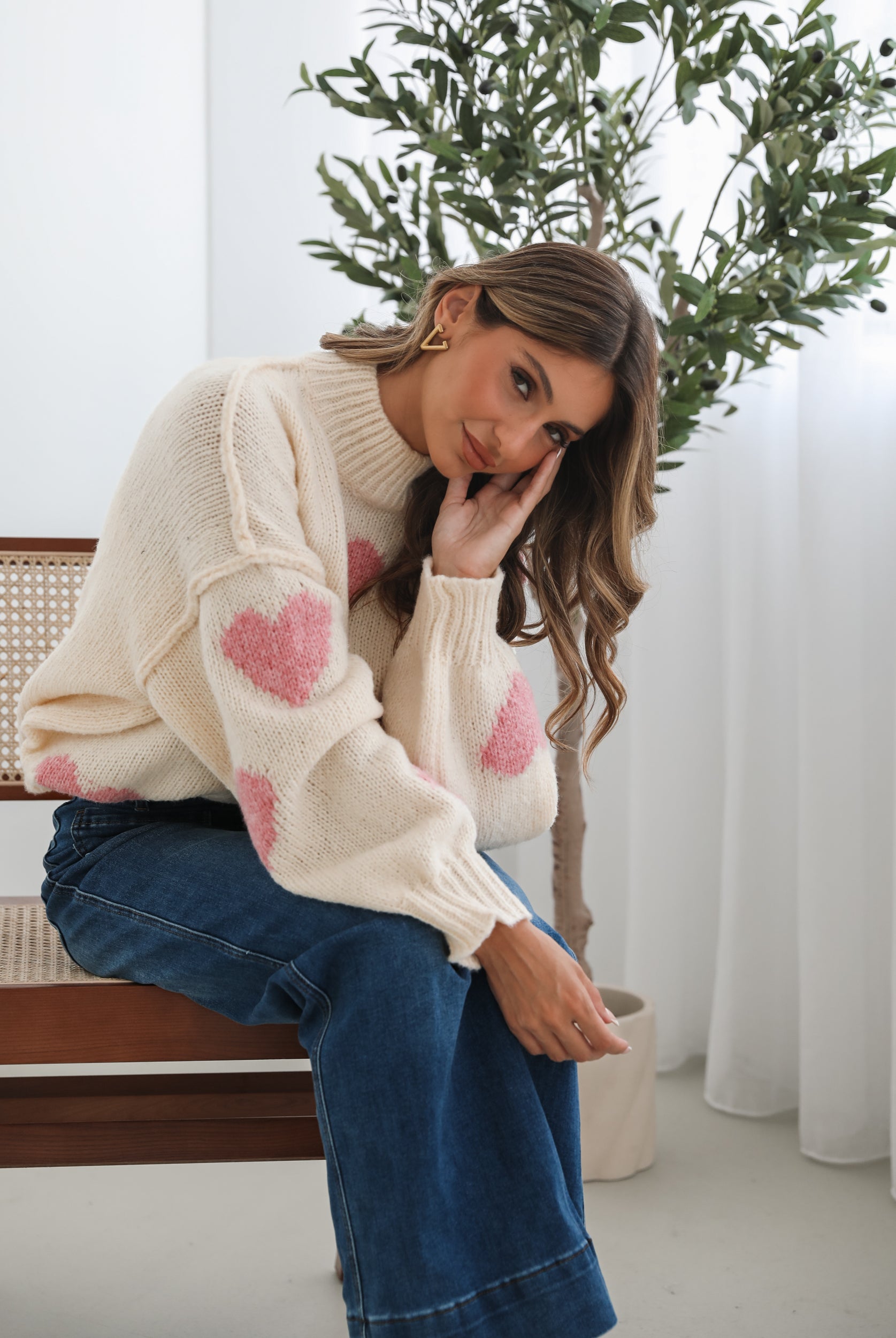 A woman sits smiling on a wooden chair with a woven back and seat. She is wearing a cozy cream-colored sweater with pink heart patterns and dark blue wide-leg jeans. Her long, wavy hair is styled to one side, and she accessorizes with gold triangle-shaped earrings. A tall green leafy plant stands in the background against white curtains, adding a fresh, modern touch to the bright, minimal setting