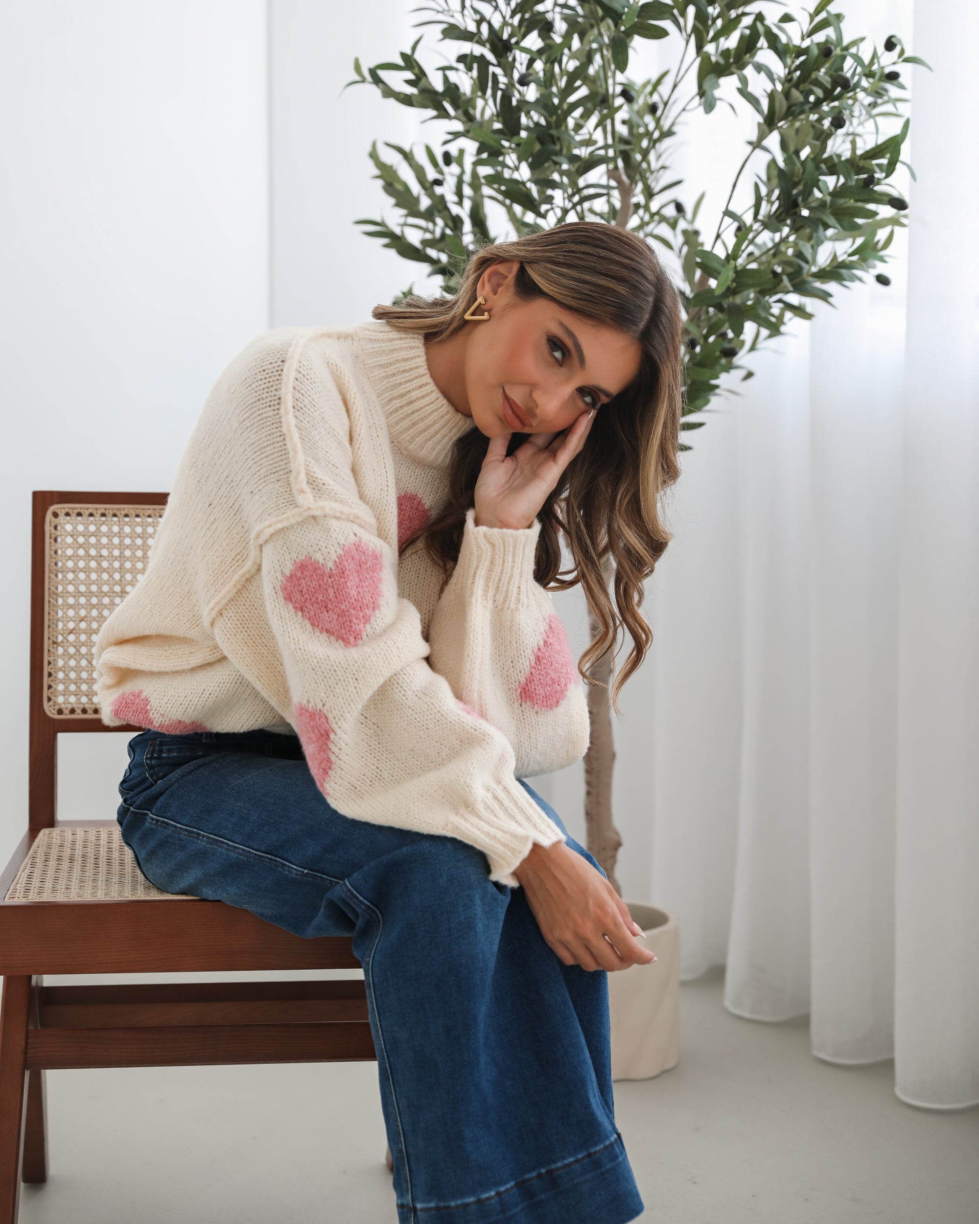 A woman sits smiling on a wooden chair with a woven back and seat. She is wearing a cozy cream-colored sweater with pink heart patterns and dark blue wide-leg jeans. Her long, wavy hair is styled to one side, and she accessorizes with gold triangle-shaped earrings. A tall green leafy plant stands in the background against white curtains, adding a fresh, modern touch to the bright, minimal setting