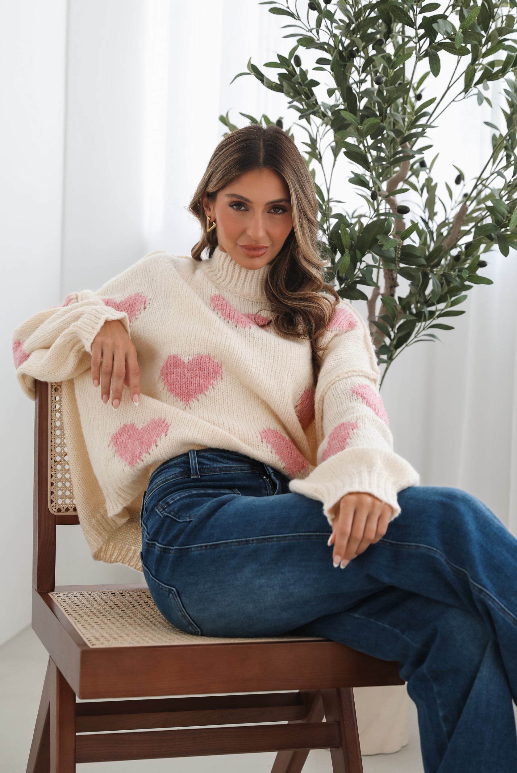 A woman sits smiling on a wooden chair with a woven back and seat. She is wearing a cozy cream-colored sweater with pink heart patterns and dark blue wide-leg jeans. Her long, wavy hair is styled to one side, and she accessorizes with gold triangle-shaped earrings. A tall green leafy plant stands in the background against white curtains, adding a fresh, modern touch to the bright, minimal setting
