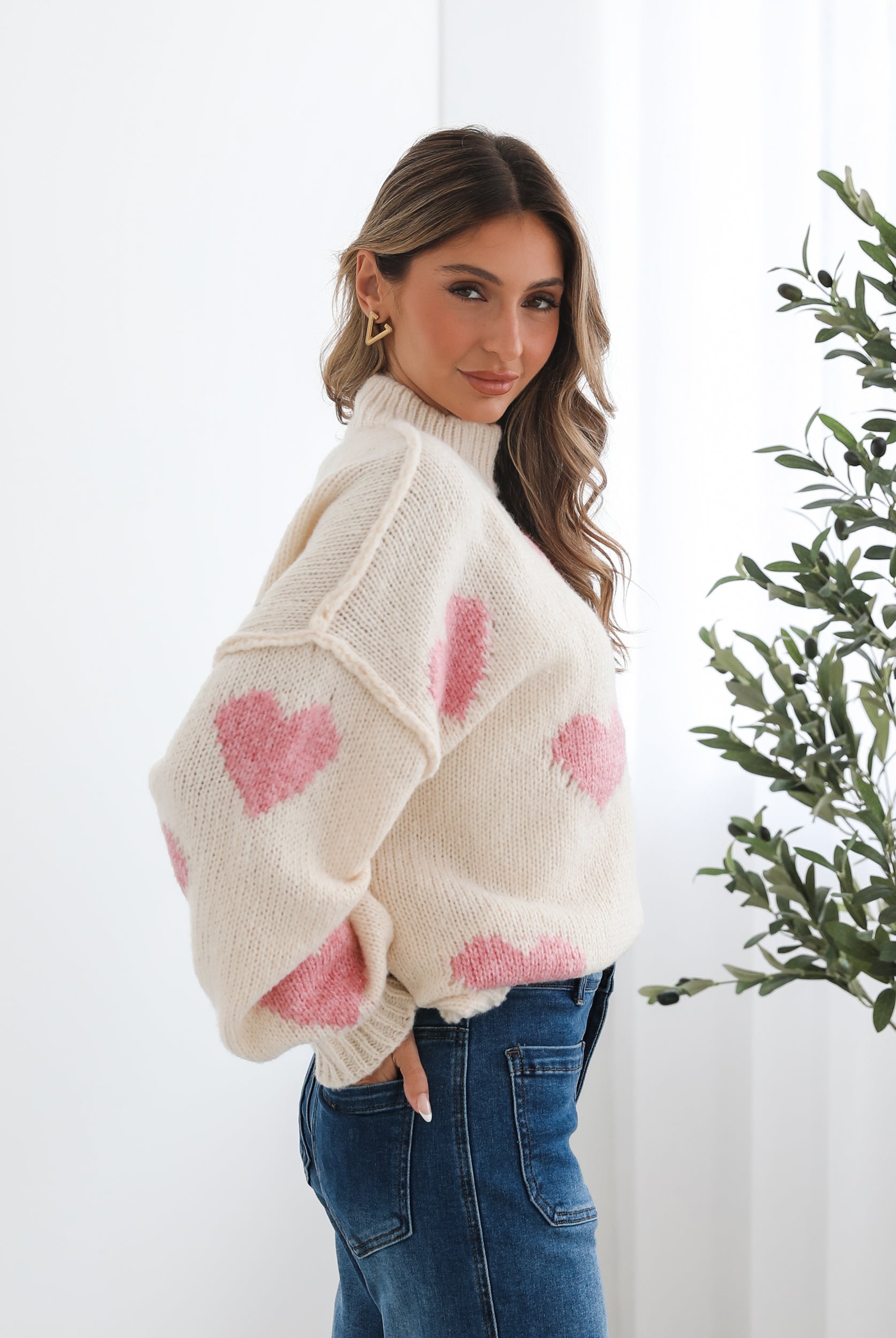 A woman sits smiling on a wooden chair with a woven back and seat. She is wearing a cozy cream-colored sweater with pink heart patterns and dark blue wide-leg jeans. Her long, wavy hair is styled to one side, and she accessorizes with gold triangle-shaped earrings. A tall green leafy plant stands in the background against white curtains, adding a fresh, modern touch to the bright, minimal setting