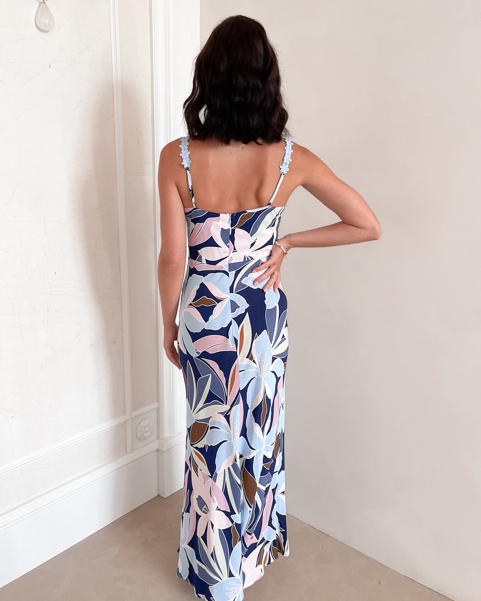 A woman stands smiling indoors against a light-colored wall, wearing a fitted, floor-length dress with a bold floral print in shades of navy blue, light blue, pink, and white. The dress features a deep V-neckline and light blue floral straps. Her dark, wavy hair is styled down, and she accessorizes with a simple gold necklace and rings. She poses confidently with her hands on her hips and is barefoot, adding to the relaxed and elegant vibe of the scene