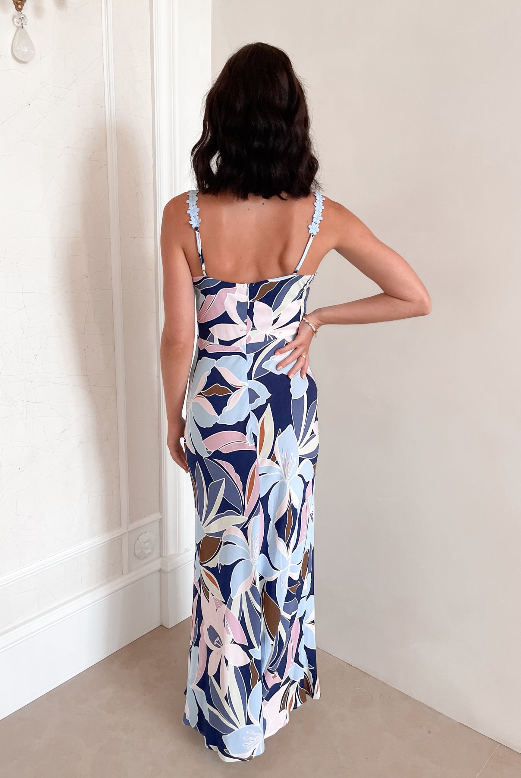 A woman stands smiling indoors against a light-colored wall, wearing a fitted, floor-length dress with a bold floral print in shades of navy blue, light blue, pink, and white. The dress features a deep V-neckline and light blue floral straps. Her dark, wavy hair is styled down, and she accessorizes with a simple gold necklace and rings. She poses confidently with her hands on her hips and is barefoot, adding to the relaxed and elegant vibe of the scene