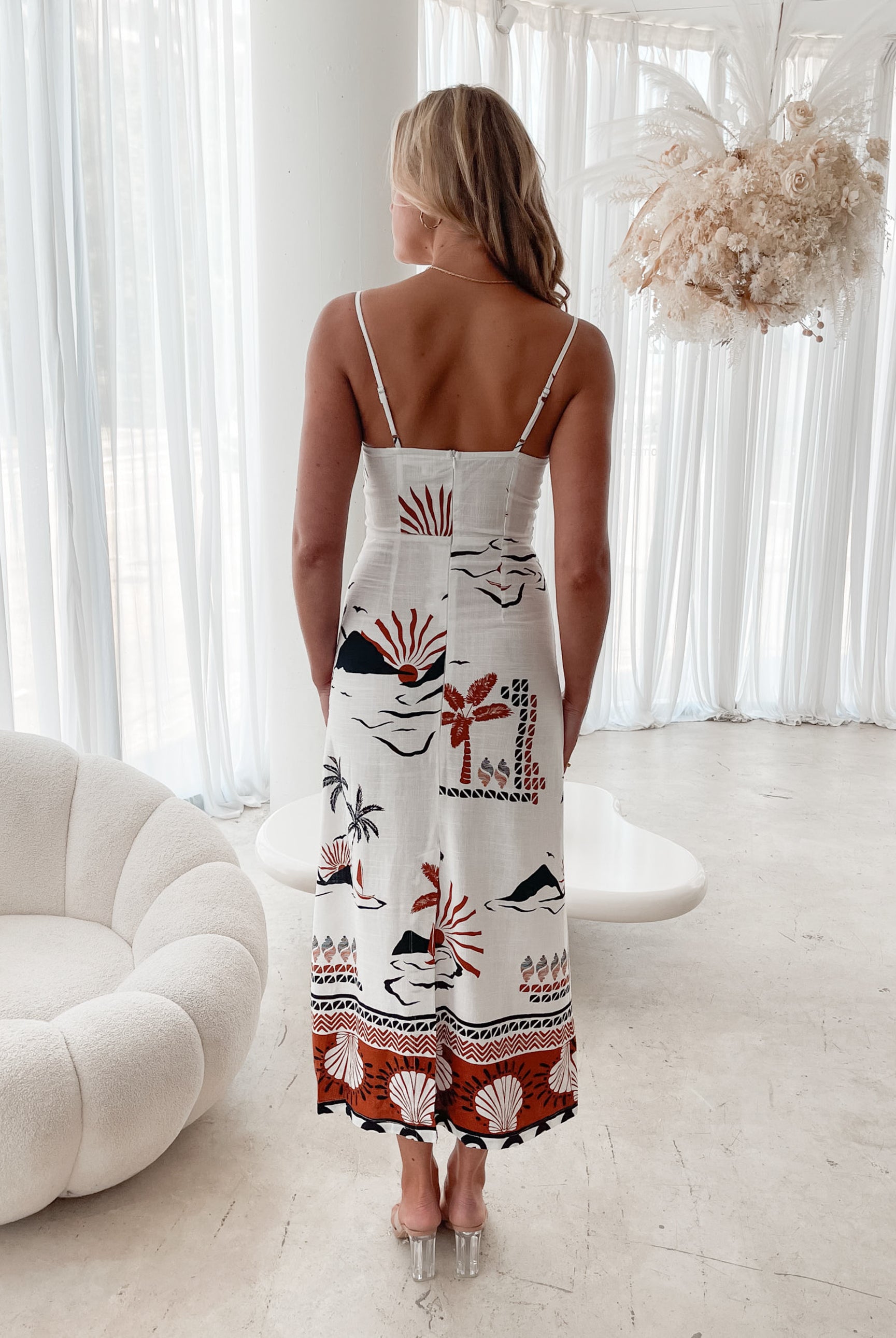 A woman stands indoors in a softly lit, modern room with white sheer curtains and neutral-toned decor, including a round textured chair and a sculptural coffee table. She is wearing a white, fitted, spaghetti-strap sundress featuring a tropical print with dolphins, palm trees, and sunset motifs in black, red, and orange tones. She has wavy blonde hair, nude heels, and a delicate necklace, exuding a relaxed, summery vibe.