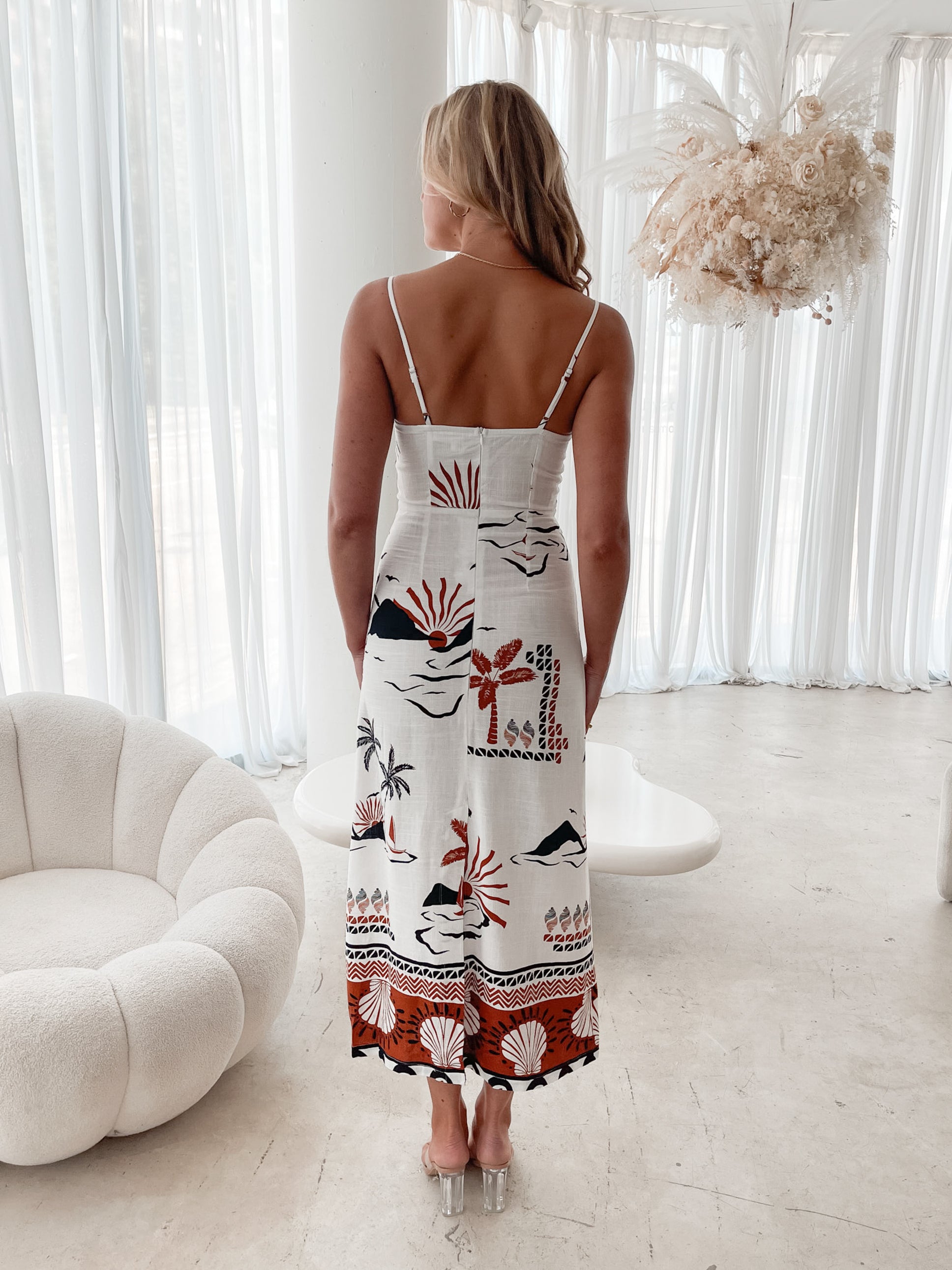 A woman stands indoors in a softly lit, modern room with white sheer curtains and neutral-toned decor, including a round textured chair and a sculptural coffee table. She is wearing a white, fitted, spaghetti-strap sundress featuring a tropical print with dolphins, palm trees, and sunset motifs in black, red, and orange tones. She has wavy blonde hair, nude heels, and a delicate necklace, exuding a relaxed, summery vibe.