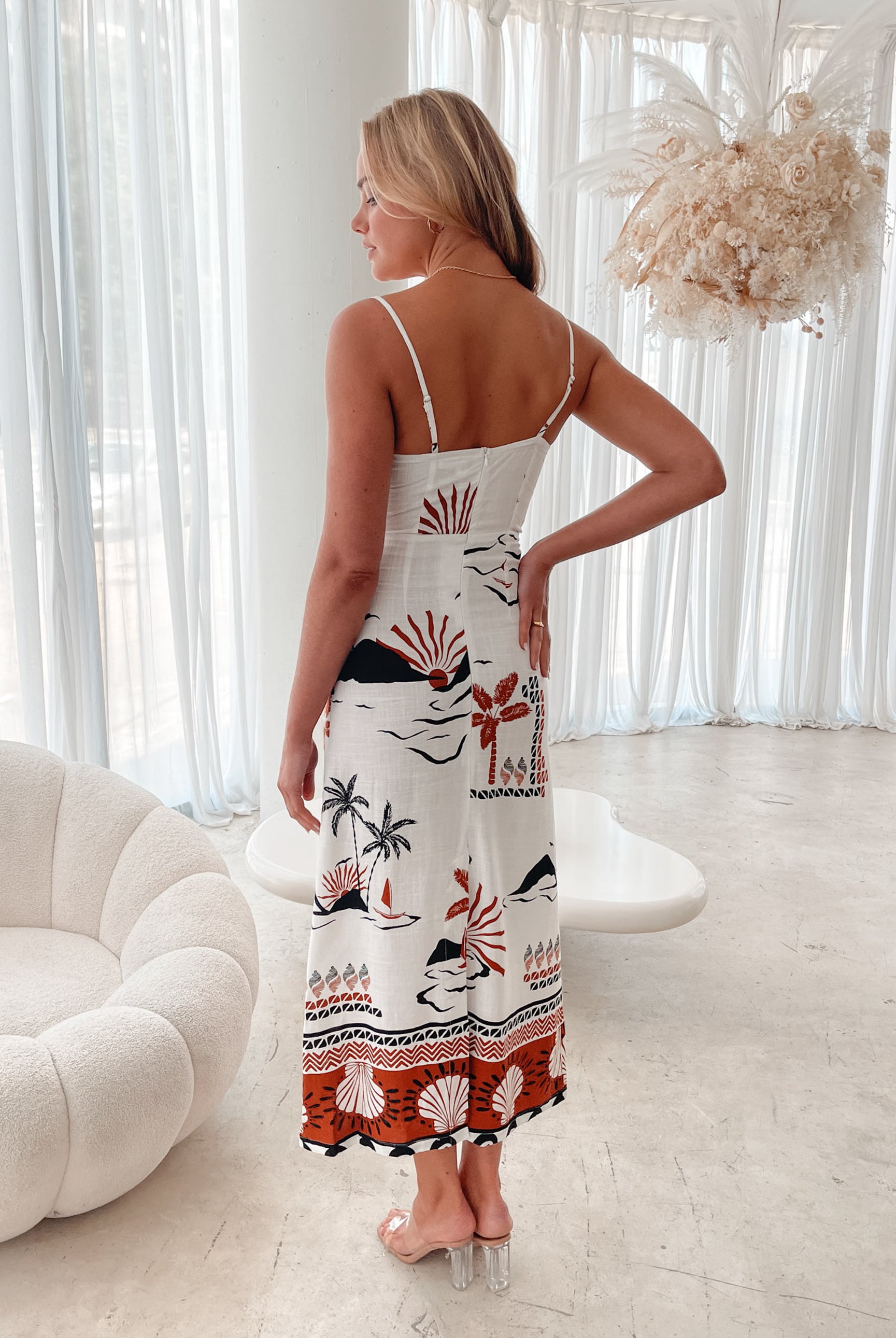 A woman stands indoors in a softly lit, modern room with white sheer curtains and neutral-toned decor, including a round textured chair and a sculptural coffee table. She is wearing a white, fitted, spaghetti-strap sundress featuring a tropical print with dolphins, palm trees, and sunset motifs in black, red, and orange tones. She has wavy blonde hair, nude heels, and a delicate necklace, exuding a relaxed, summery vibe.