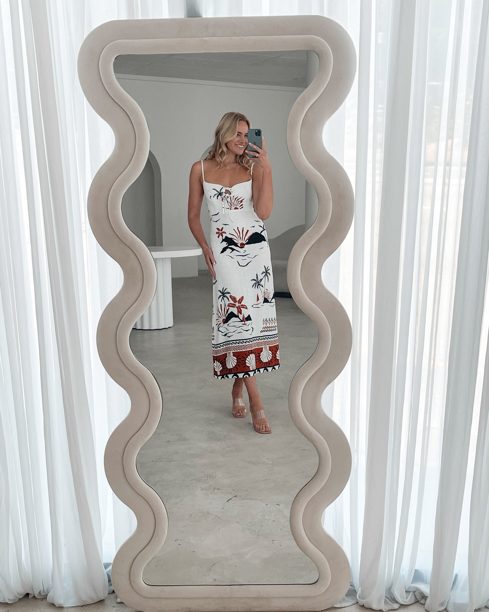 A woman stands indoors in a softly lit, modern room with white sheer curtains and neutral-toned decor, including a round textured chair and a sculptural coffee table. She is wearing a white, fitted, spaghetti-strap sundress featuring a tropical print with dolphins, palm trees, and sunset motifs in black, red, and orange tones. She has wavy blonde hair, nude heels, and a delicate necklace, exuding a relaxed, summery vibe.
