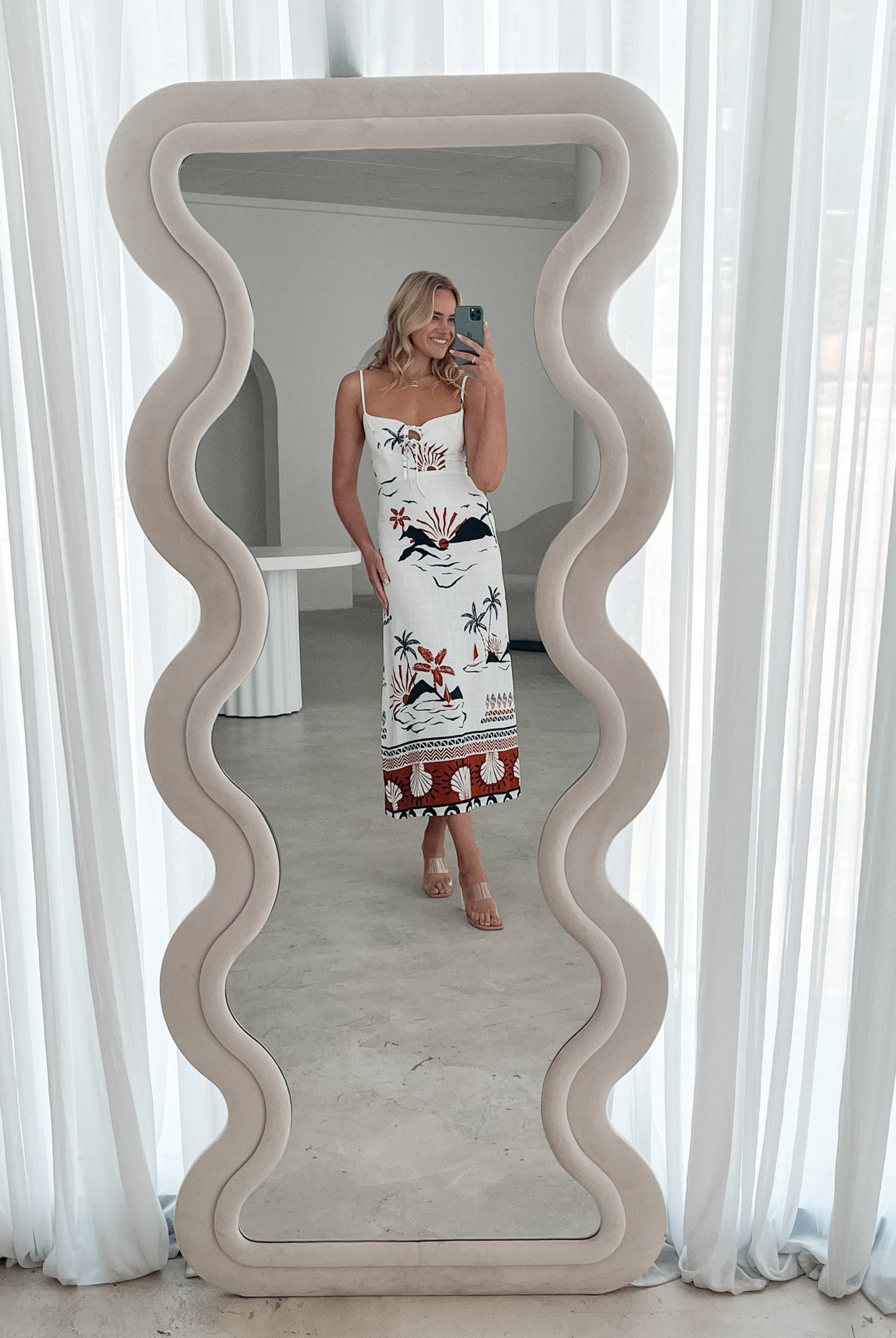 A woman stands indoors in a softly lit, modern room with white sheer curtains and neutral-toned decor, including a round textured chair and a sculptural coffee table. She is wearing a white, fitted, spaghetti-strap sundress featuring a tropical print with dolphins, palm trees, and sunset motifs in black, red, and orange tones. She has wavy blonde hair, nude heels, and a delicate necklace, exuding a relaxed, summery vibe.