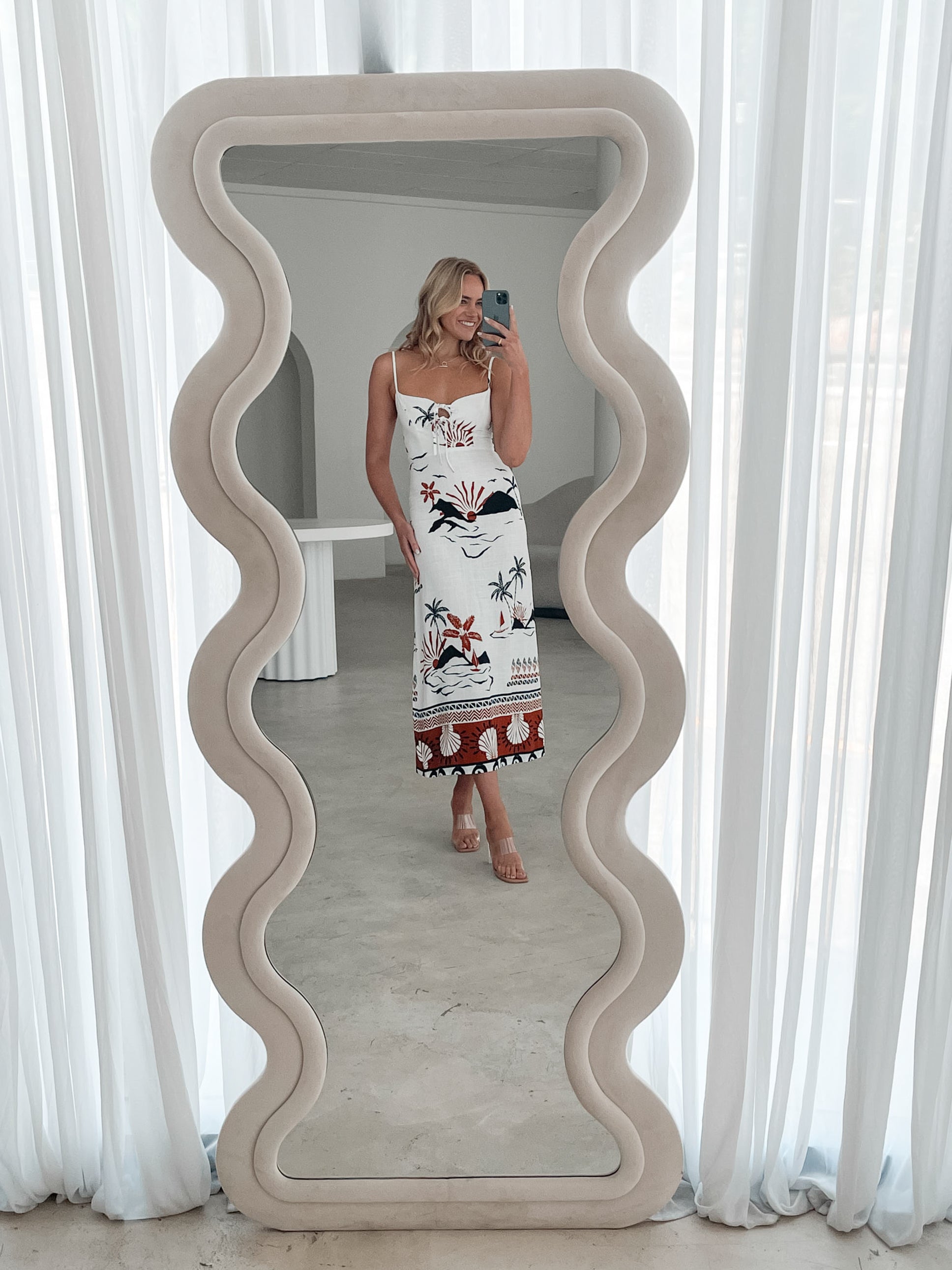 A woman stands indoors in a softly lit, modern room with white sheer curtains and neutral-toned decor, including a round textured chair and a sculptural coffee table. She is wearing a white, fitted, spaghetti-strap sundress featuring a tropical print with dolphins, palm trees, and sunset motifs in black, red, and orange tones. She has wavy blonde hair, nude heels, and a delicate necklace, exuding a relaxed, summery vibe.