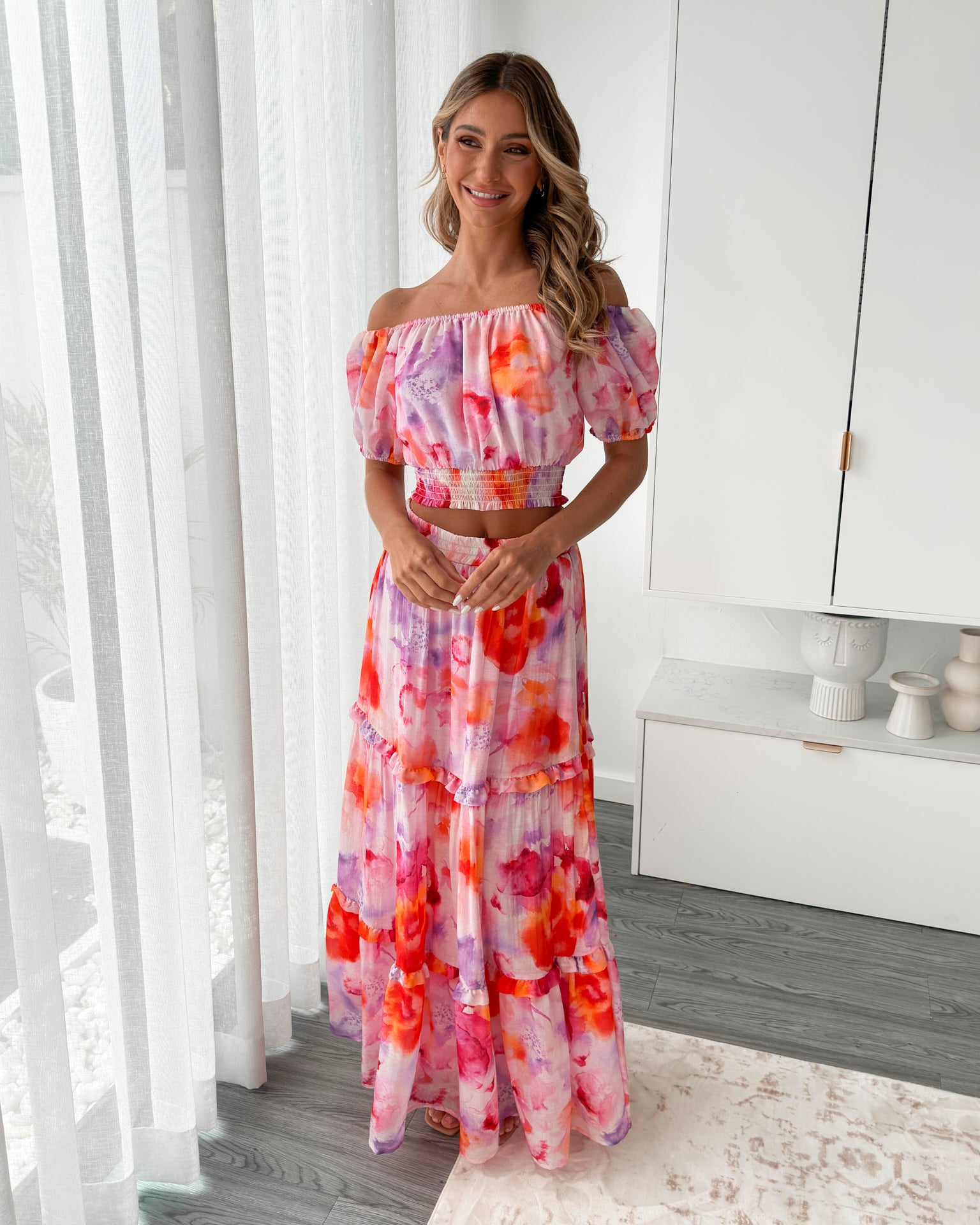 A woman stands in a bright, modern room with sheer curtains and minimalist white cabinetry. She is wearing an off-the-shoulder, two-piece maxi outfit with a ruffled top and high-waisted skirt featuring a vibrant watercolor floral print in shades of pink, purple, orange, and red. Her long wavy hair cascades over her shoulders, and she gently holds a part of her flowing skirt, creating a graceful, summery look.