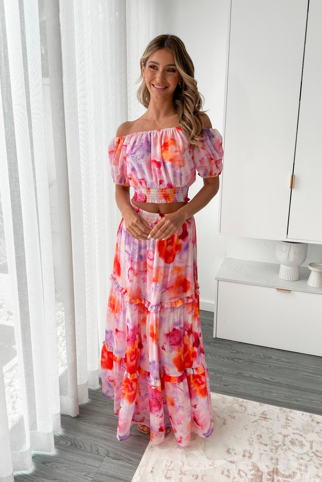 A woman stands in a bright, modern room with sheer curtains and minimalist white cabinetry. She is wearing an off-the-shoulder, two-piece maxi outfit with a ruffled top and high-waisted skirt featuring a vibrant watercolor floral print in shades of pink, purple, orange, and red. Her long wavy hair cascades over her shoulders, and she gently holds a part of her flowing skirt, creating a graceful, summery look.