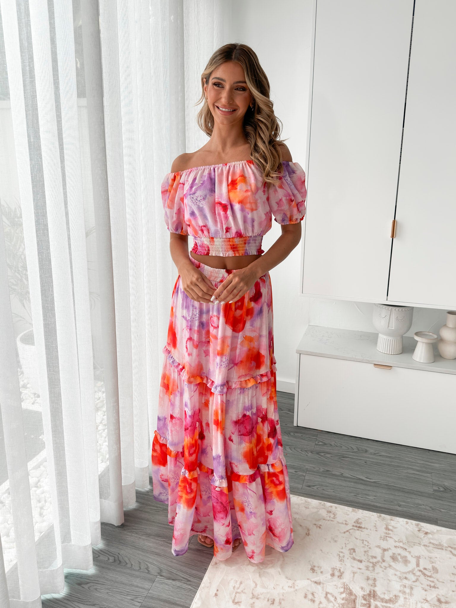 A woman stands in a bright, modern room with sheer curtains and minimalist white cabinetry. She is wearing an off-the-shoulder, two-piece maxi outfit with a ruffled top and high-waisted skirt featuring a vibrant watercolor floral print in shades of pink, purple, orange, and red. Her long wavy hair cascades over her shoulders, and she gently holds a part of her flowing skirt, creating a graceful, summery look.