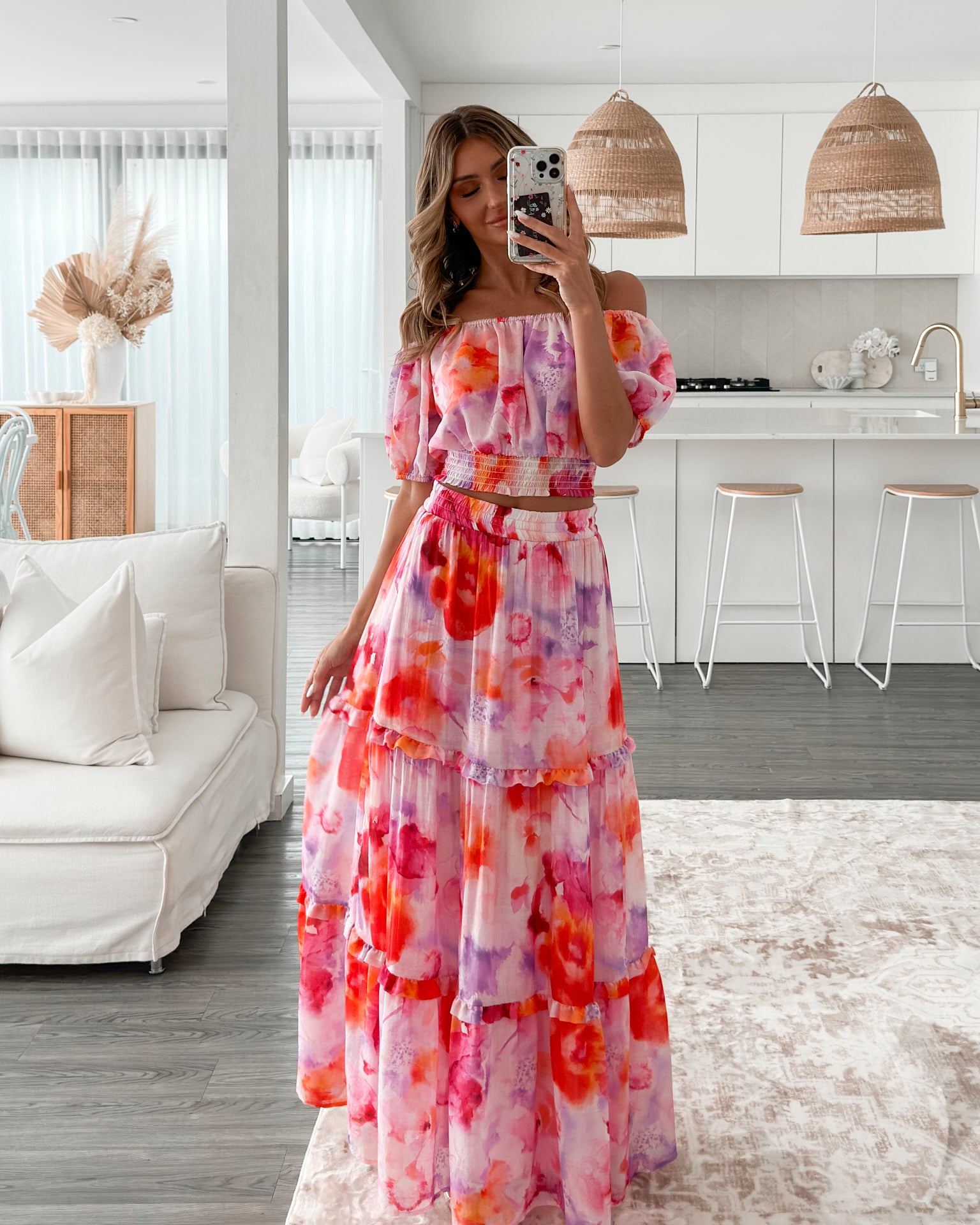 A woman stands in a bright, modern room with sheer curtains and minimalist white cabinetry. She is wearing an off-the-shoulder, two-piece maxi outfit with a ruffled top and high-waisted skirt featuring a vibrant watercolor floral print in shades of pink, purple, orange, and red. Her long wavy hair cascades over her shoulders, and she gently holds a part of her flowing skirt, creating a graceful, summery look.