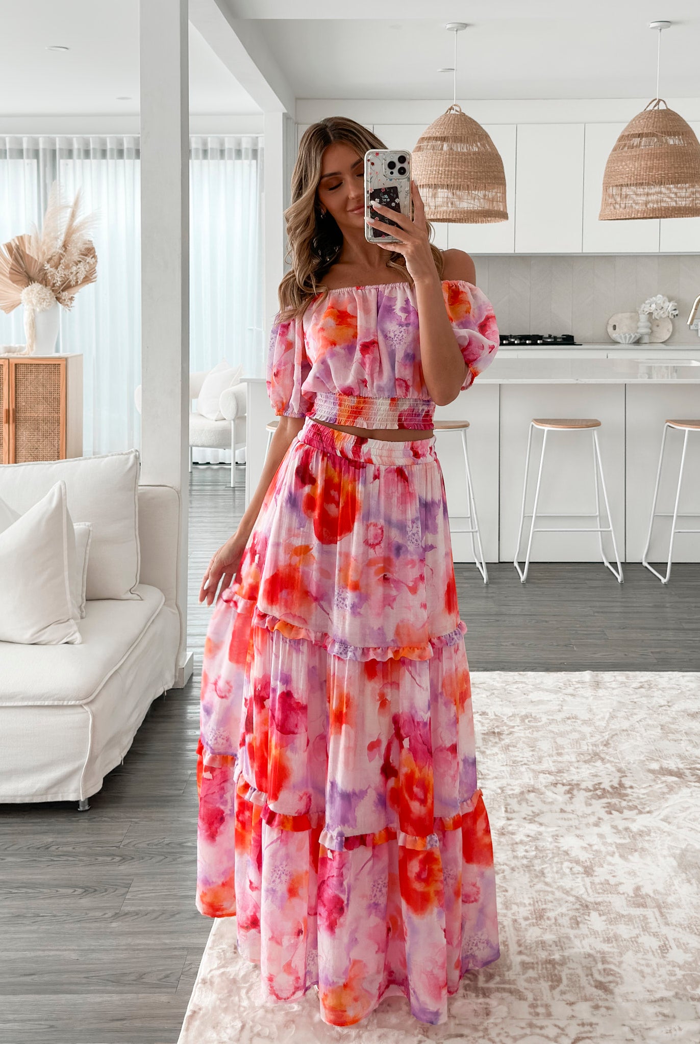 A woman stands in a bright, modern room with sheer curtains and minimalist white cabinetry. She is wearing an off-the-shoulder, two-piece maxi outfit with a ruffled top and high-waisted skirt featuring a vibrant watercolor floral print in shades of pink, purple, orange, and red. Her long wavy hair cascades over her shoulders, and she gently holds a part of her flowing skirt, creating a graceful, summery look.