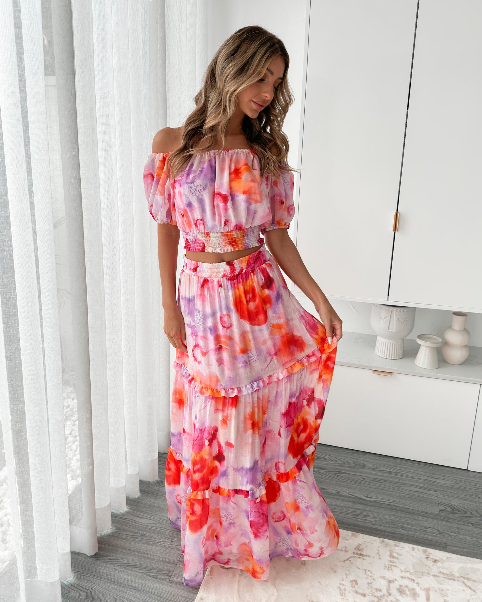 A woman stands in a bright, modern room with sheer curtains and minimalist white cabinetry. She is wearing an off-the-shoulder, two-piece maxi outfit with a ruffled top and high-waisted skirt featuring a vibrant watercolor floral print in shades of pink, purple, orange, and red. Her long wavy hair cascades over her shoulders, and she gently holds a part of her flowing skirt, creating a graceful, summery look.