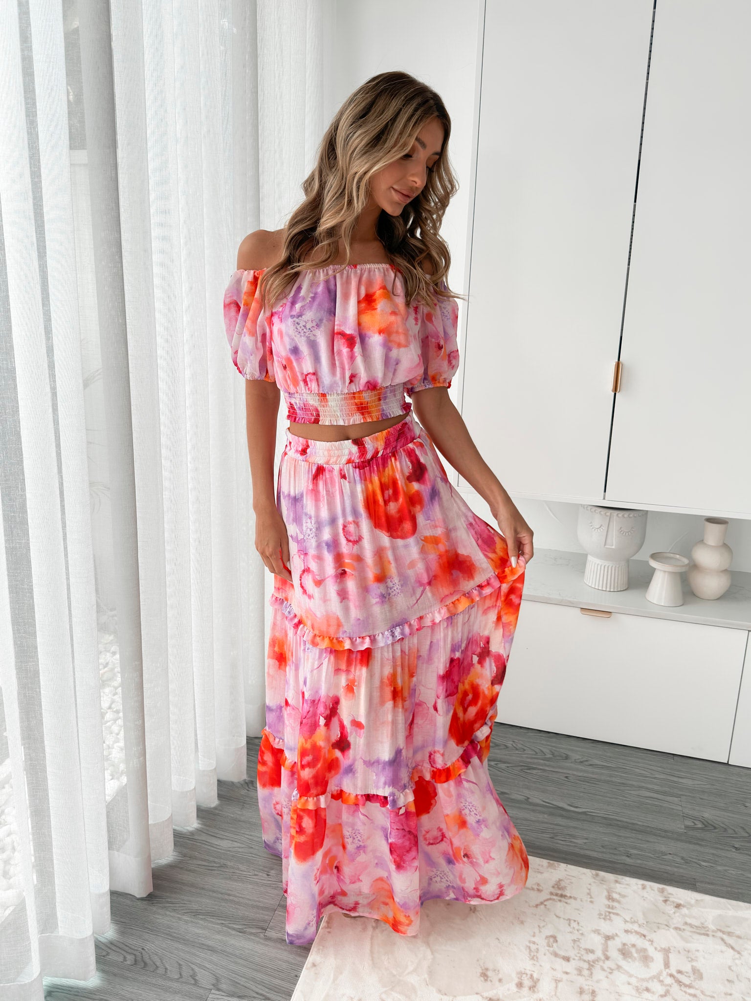 A woman stands in a bright, modern room with sheer curtains and minimalist white cabinetry. She is wearing an off-the-shoulder, two-piece maxi outfit with a ruffled top and high-waisted skirt featuring a vibrant watercolor floral print in shades of pink, purple, orange, and red. Her long wavy hair cascades over her shoulders, and she gently holds a part of her flowing skirt, creating a graceful, summery look.
