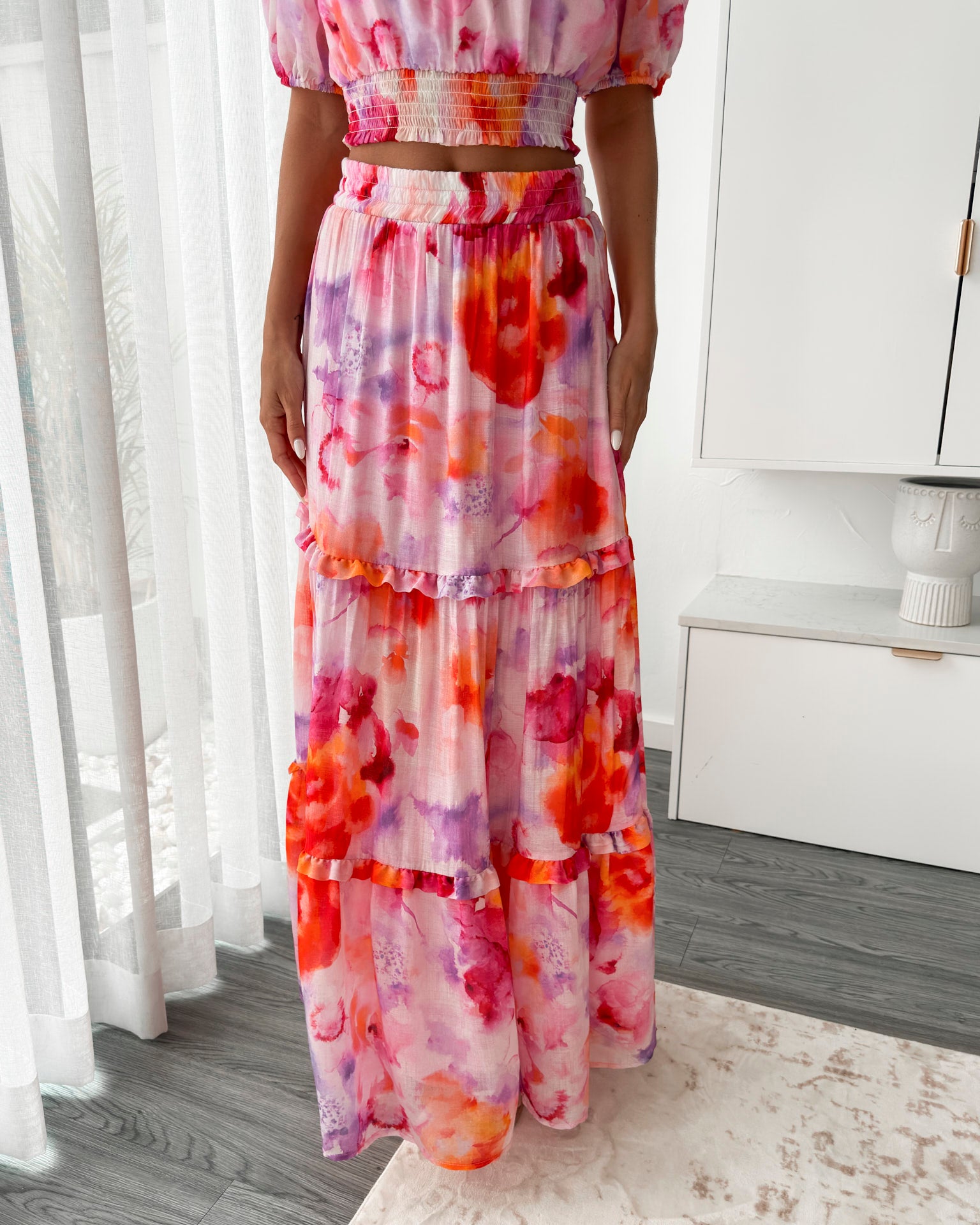 A woman stands in a bright, modern room with sheer curtains and minimalist white cabinetry. She is wearing an off-the-shoulder, two-piece maxi outfit with a ruffled top and high-waisted skirt featuring a vibrant watercolor floral print in shades of pink, purple, orange, and red. Her long wavy hair cascades over her shoulders, and she gently holds a part of her flowing skirt, creating a graceful, summery look.