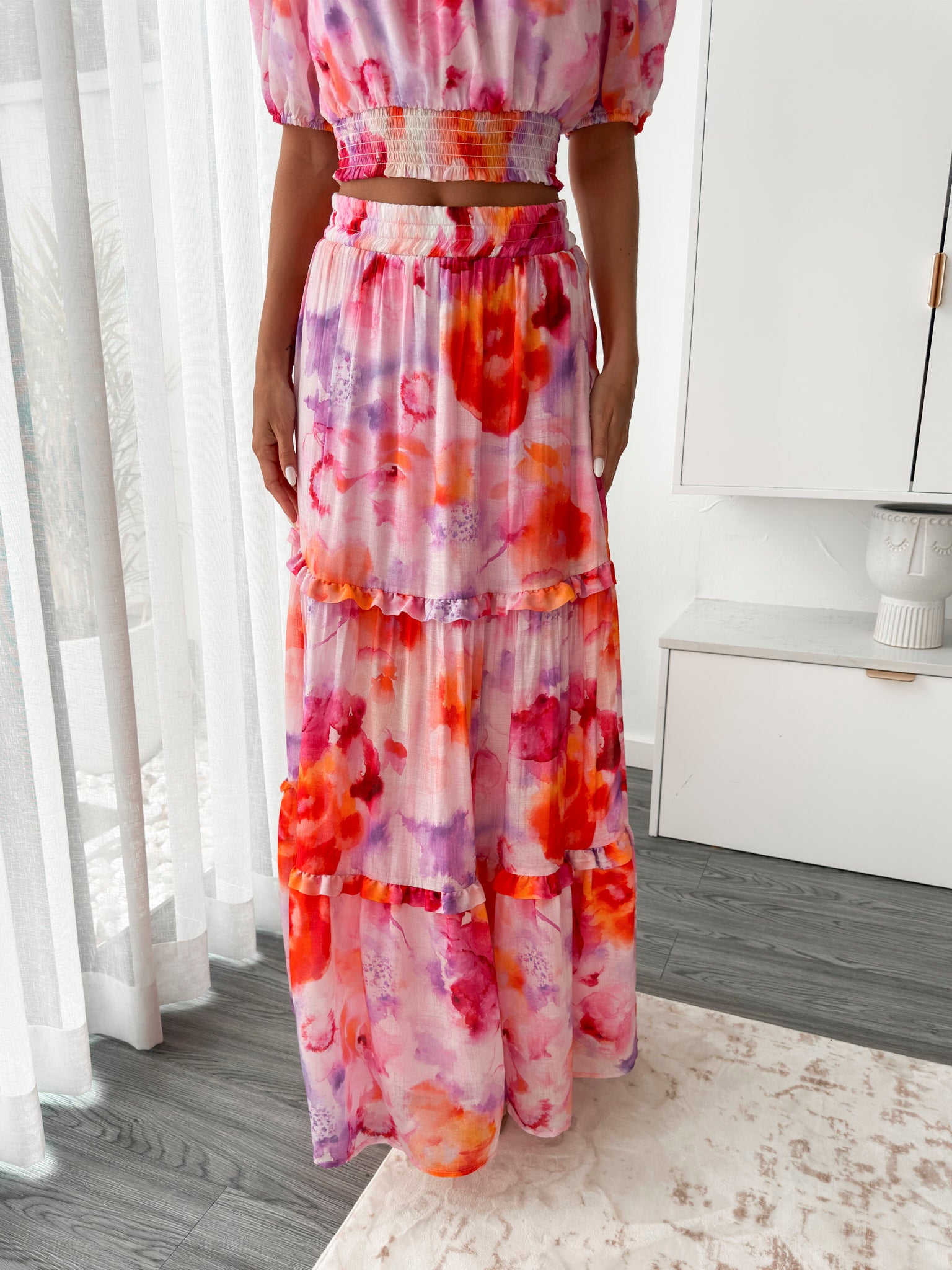 A woman stands in a bright, modern room with sheer curtains and minimalist white cabinetry. She is wearing an off-the-shoulder, two-piece maxi outfit with a ruffled top and high-waisted skirt featuring a vibrant watercolor floral print in shades of pink, purple, orange, and red. Her long wavy hair cascades over her shoulders, and she gently holds a part of her flowing skirt, creating a graceful, summery look.