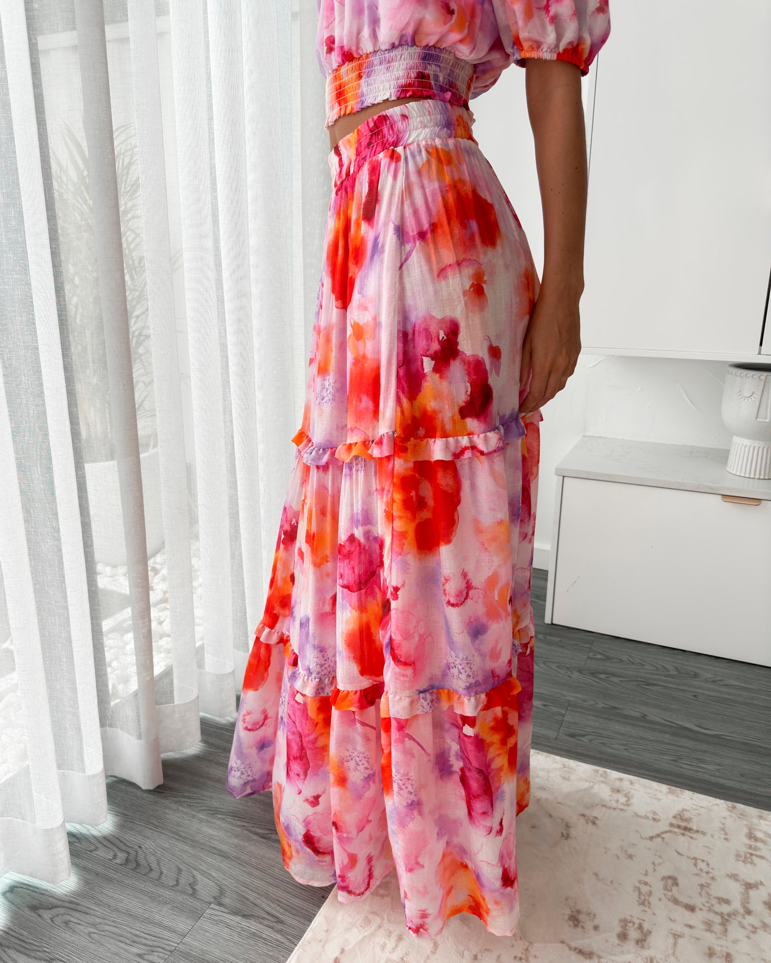 A woman stands in a bright, modern room with sheer curtains and minimalist white cabinetry. She is wearing an off-the-shoulder, two-piece maxi outfit with a ruffled top and high-waisted skirt featuring a vibrant watercolor floral print in shades of pink, purple, orange, and red. Her long wavy hair cascades over her shoulders, and she gently holds a part of her flowing skirt, creating a graceful, summery look.
