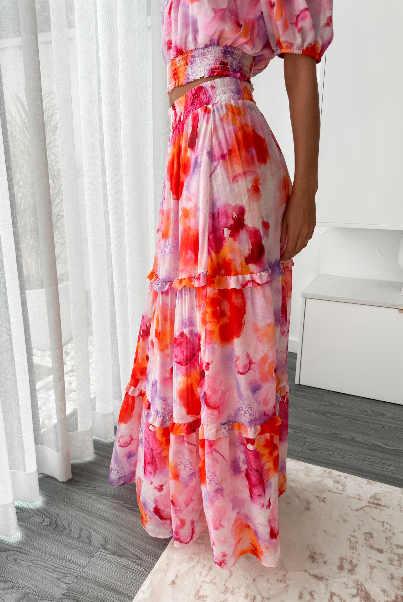 A woman stands in a bright, modern room with sheer curtains and minimalist white cabinetry. She is wearing an off-the-shoulder, two-piece maxi outfit with a ruffled top and high-waisted skirt featuring a vibrant watercolor floral print in shades of pink, purple, orange, and red. Her long wavy hair cascades over her shoulders, and she gently holds a part of her flowing skirt, creating a graceful, summery look.