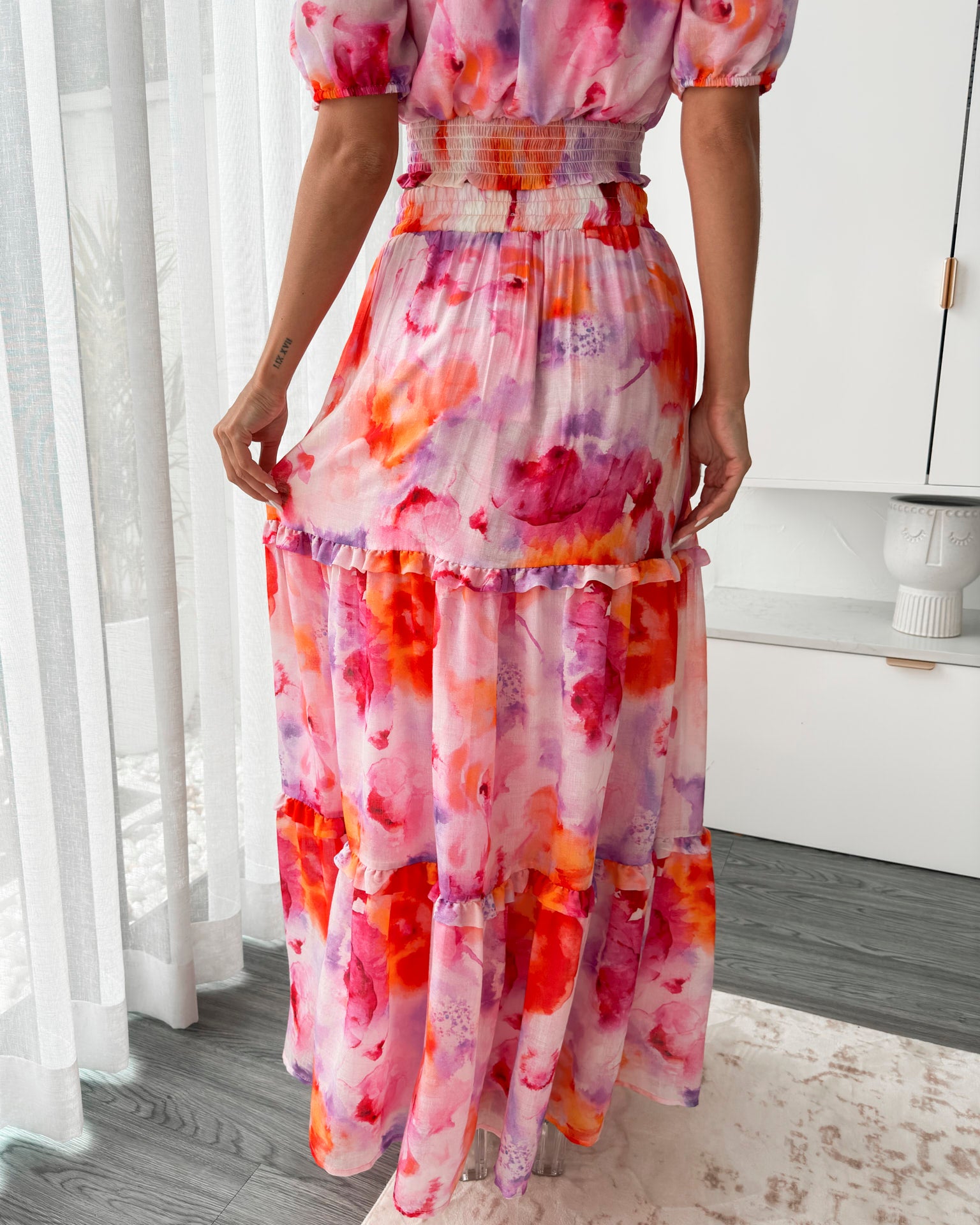A woman stands in a bright, modern room with sheer curtains and minimalist white cabinetry. She is wearing an off-the-shoulder, two-piece maxi outfit with a ruffled top and high-waisted skirt featuring a vibrant watercolor floral print in shades of pink, purple, orange, and red. Her long wavy hair cascades over her shoulders, and she gently holds a part of her flowing skirt, creating a graceful, summery look.