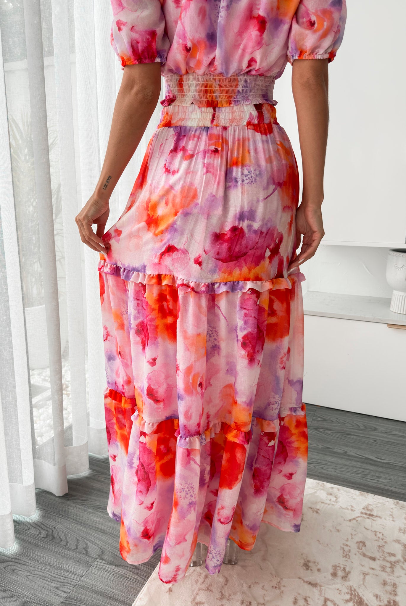 A woman stands in a bright, modern room with sheer curtains and minimalist white cabinetry. She is wearing an off-the-shoulder, two-piece maxi outfit with a ruffled top and high-waisted skirt featuring a vibrant watercolor floral print in shades of pink, purple, orange, and red. Her long wavy hair cascades over her shoulders, and she gently holds a part of her flowing skirt, creating a graceful, summery look.